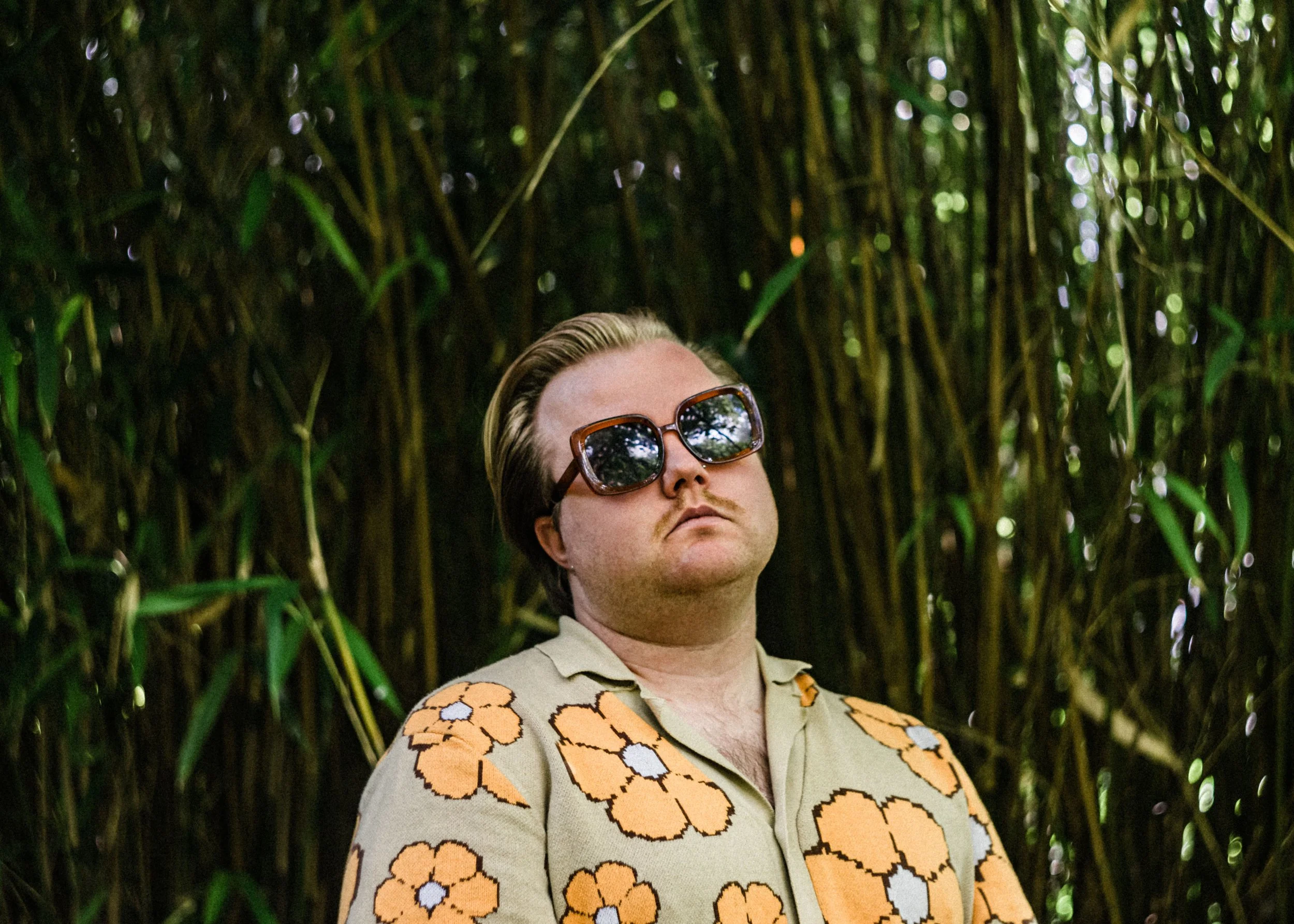 A person with short hair and sunglasses standing among tall green plants, wearing a shirt with an orange floral pattern.