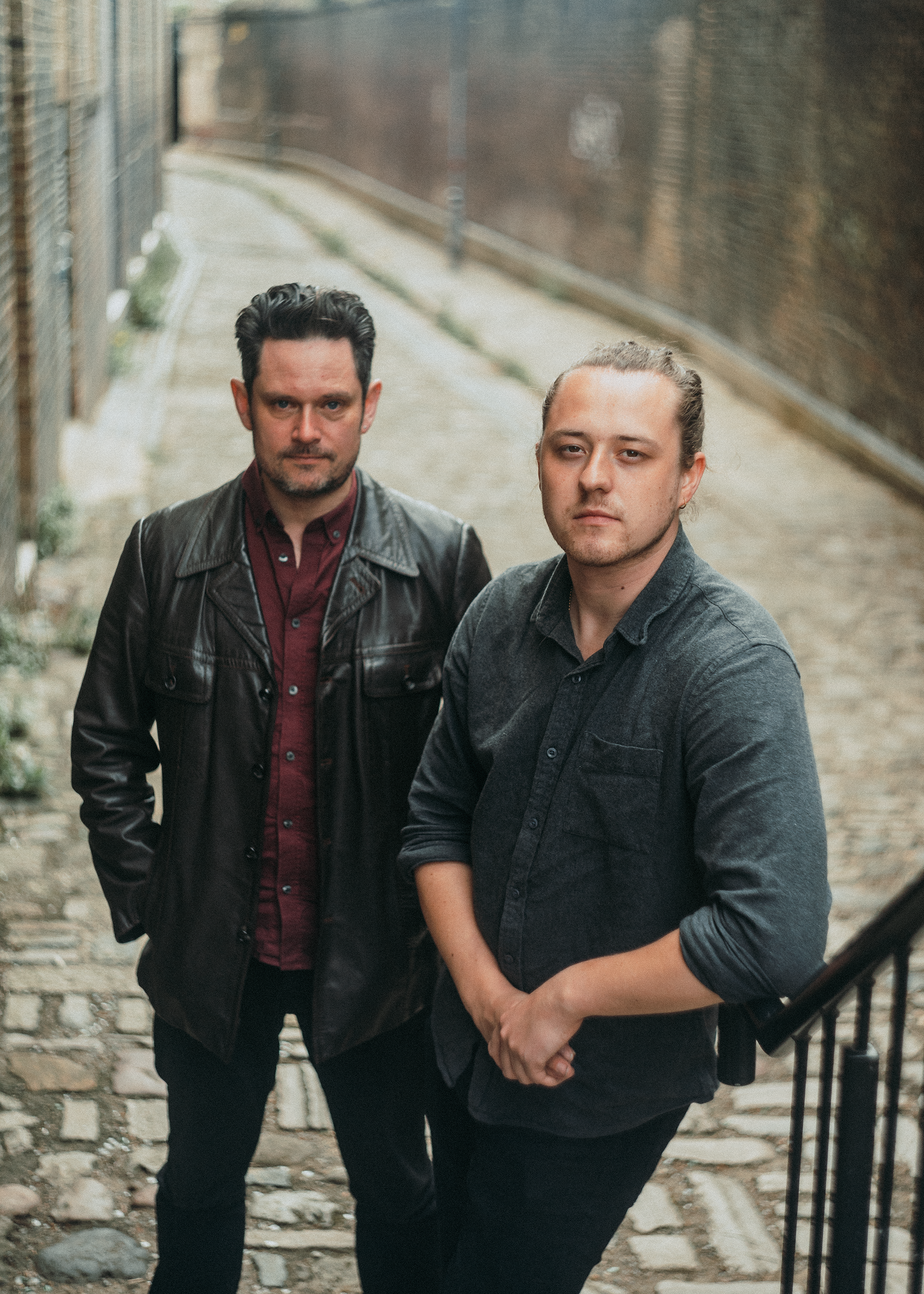 Two young men with long hair, one with a beard, stand on a cobblestone street near a brick wall, looking at the camera with serious expressions.