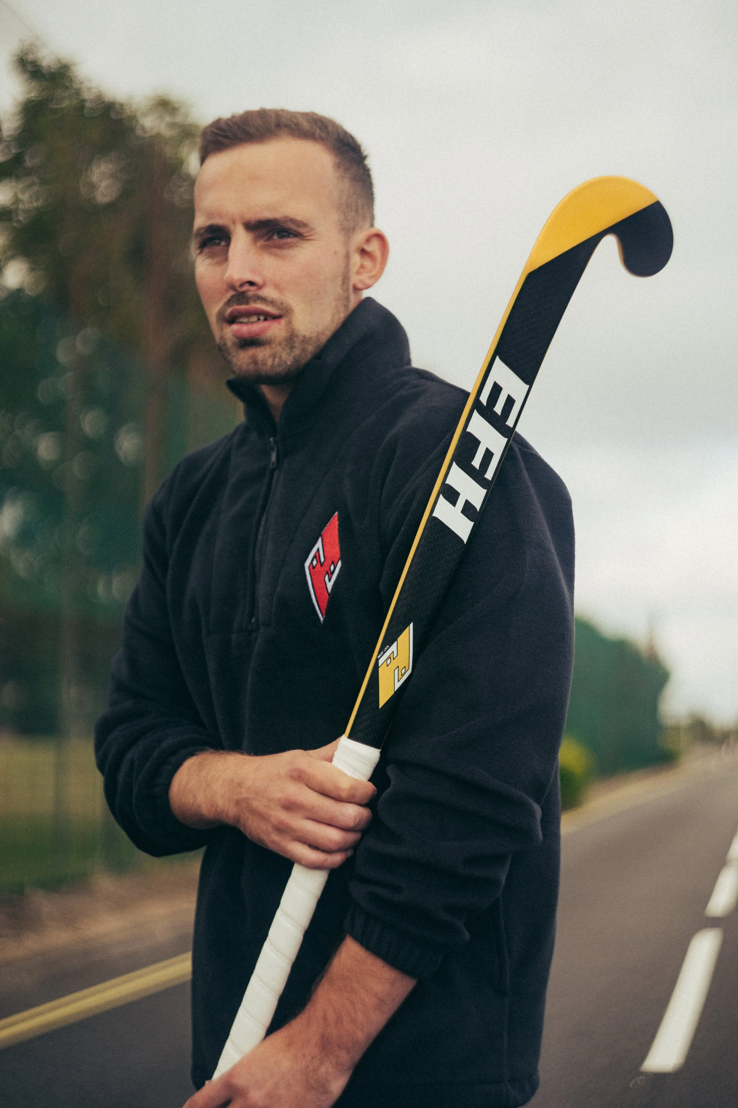 A young man holding a hockey stick outdoors, wearing a black hoodie, with a blurred background of trees and cloudy sky.