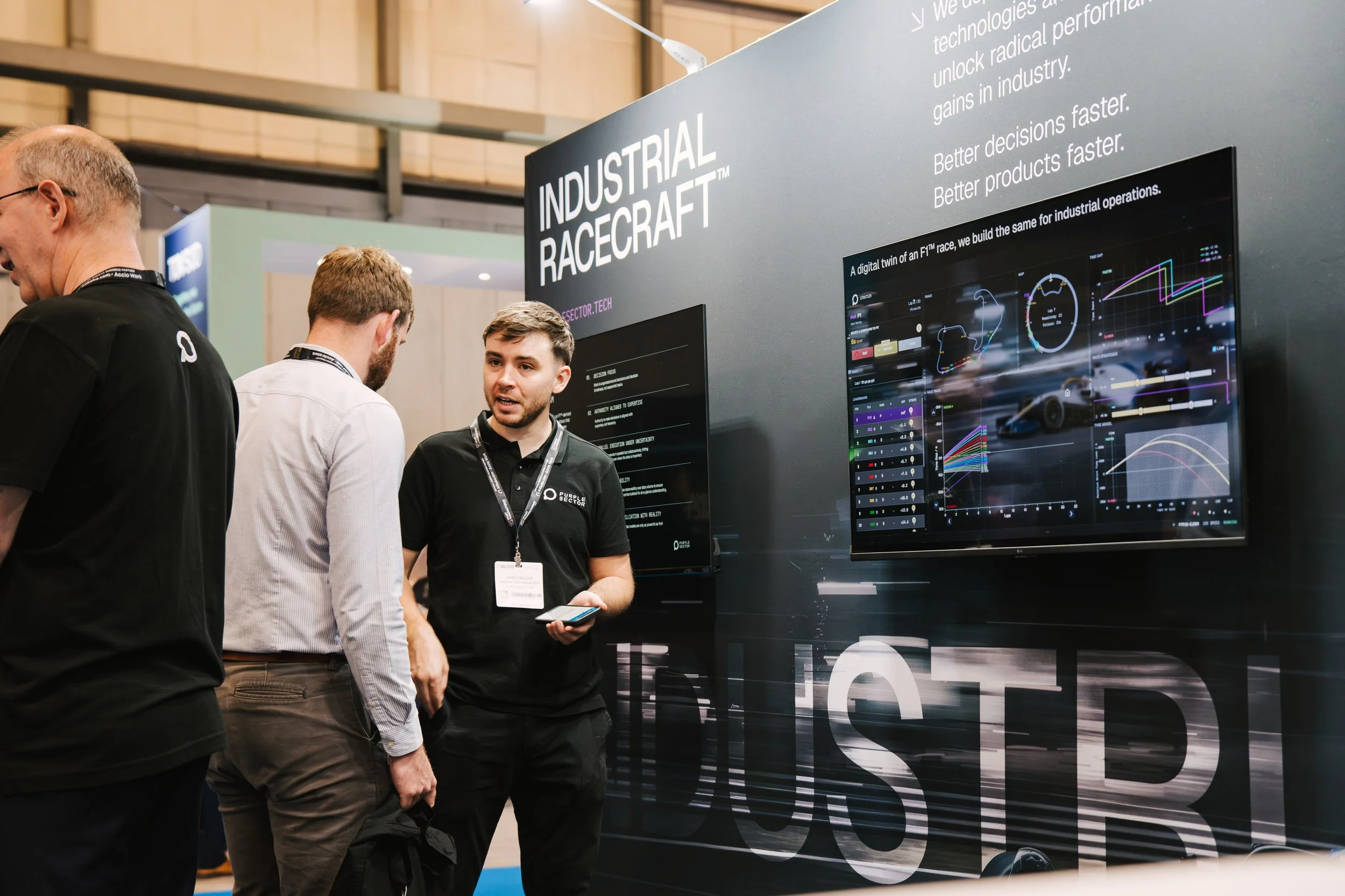 Three men are standing and talking at a trade show or exhibition booth with a large display screen showing graphs and text about industrial operations and technology. The booth has a sign saying "INDUSTRIAL RACECAR". The men are wearing conference ba