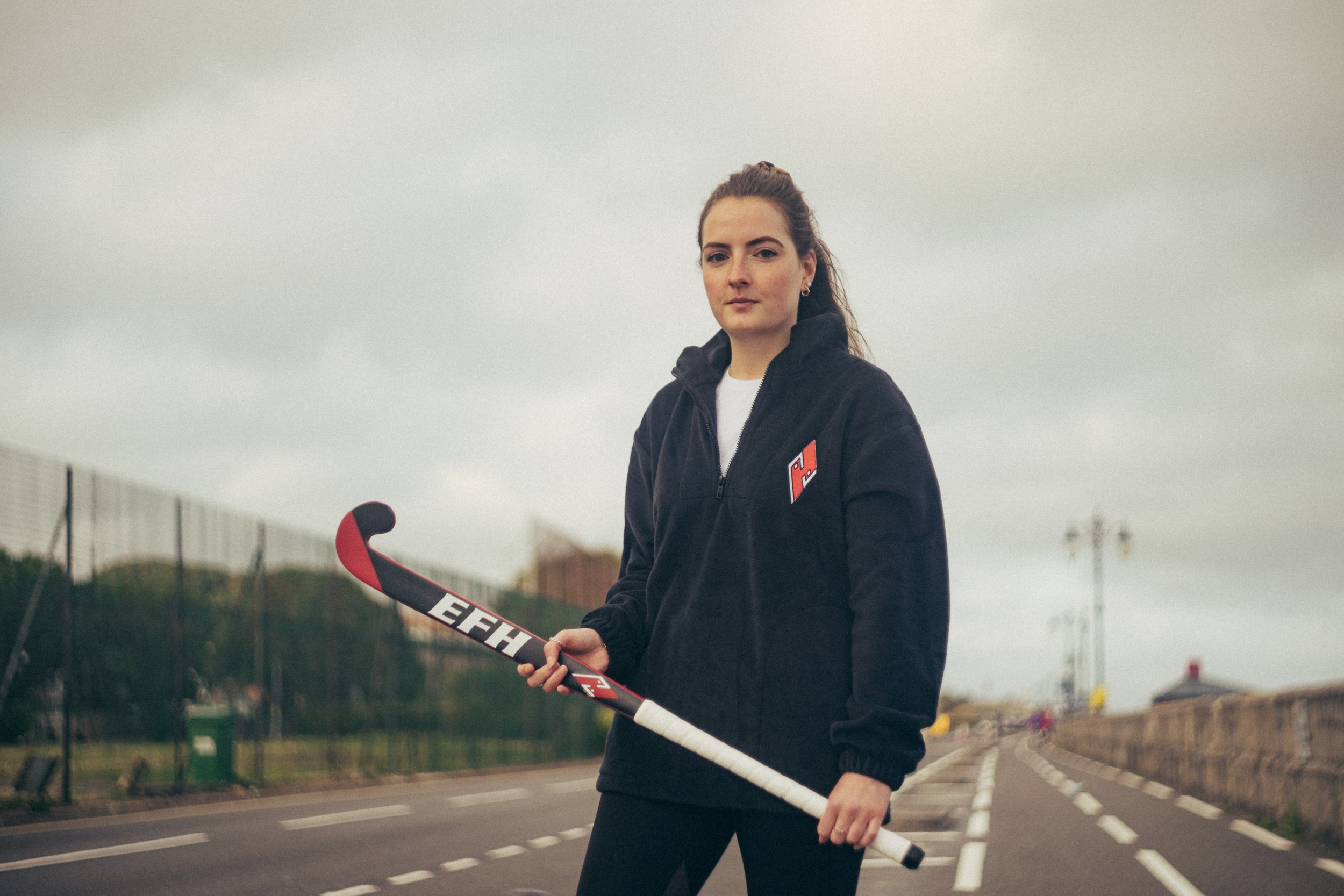 Young woman with brown hair in a ponytail holding a hockey stick outdoors on overcast day, wearing a black hoodie with a red and white logo.