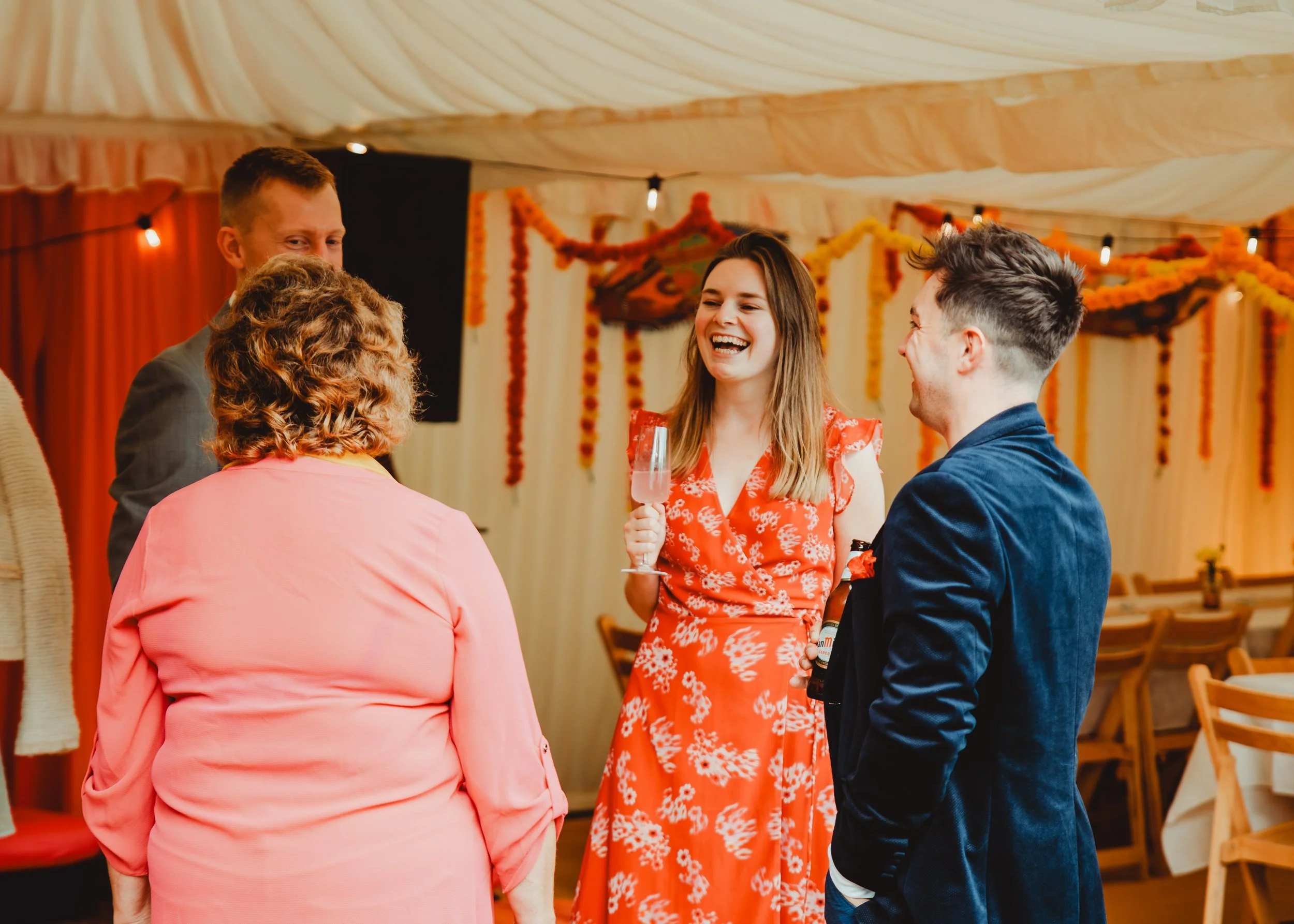 People at a social gathering indoors, including a woman in a red floral dress holding a glass of pink drink, engaged in conversation with others, decorated with marigold flower garlands and orange curtains.