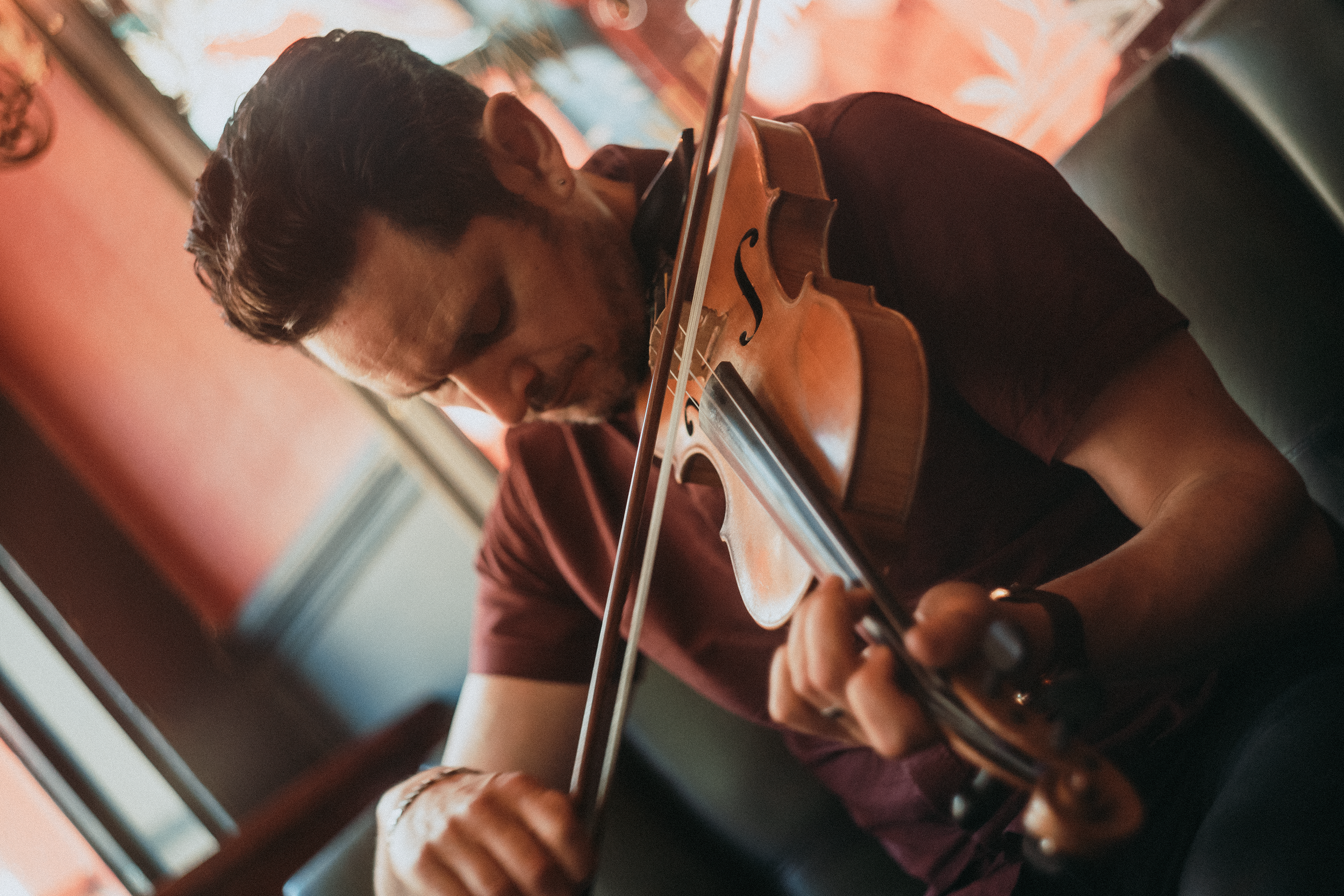 A man playing a violin, viewed from the side, in an indoor setting with warm lighting and a colorful background.
