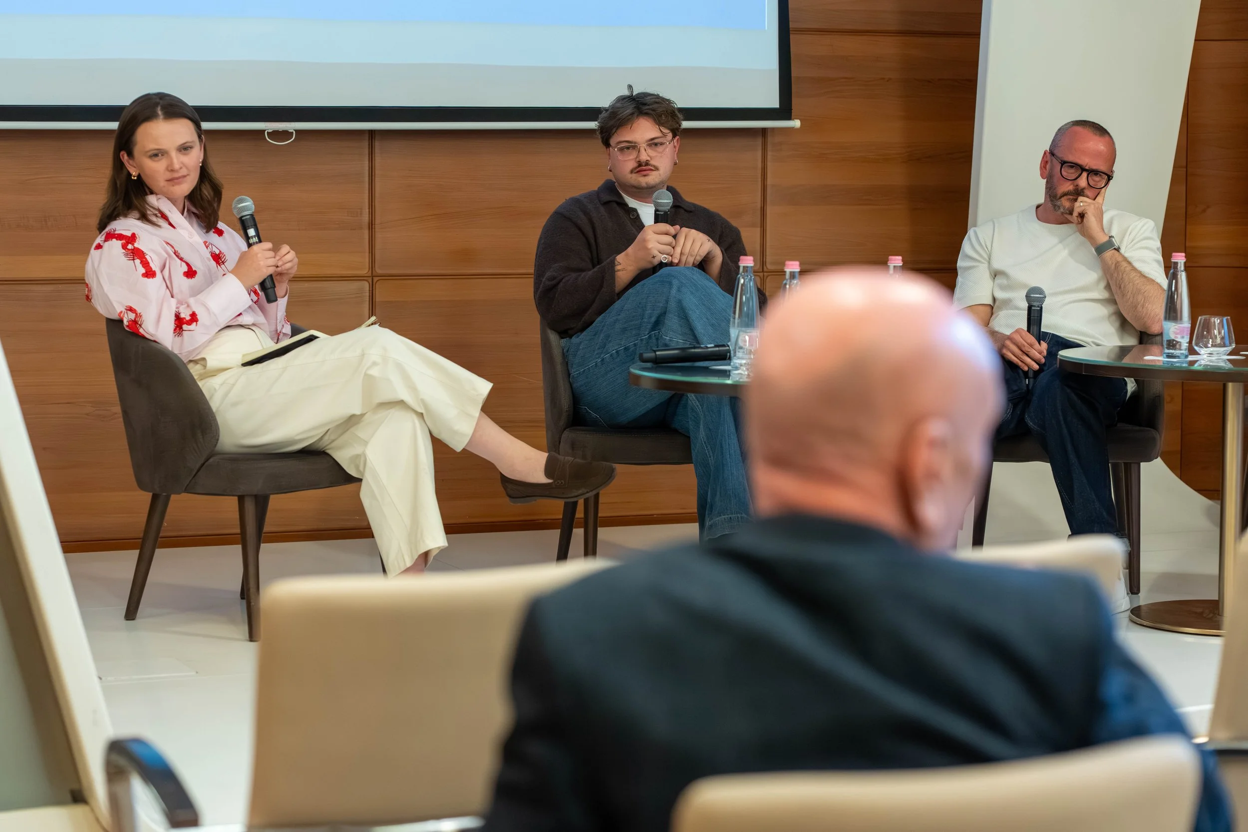 Three people sitting on stage, participating in a panel discussion, each holding a microphone, with water bottles on the table in front of them, in a room with wooden paneling and a large screen behind.