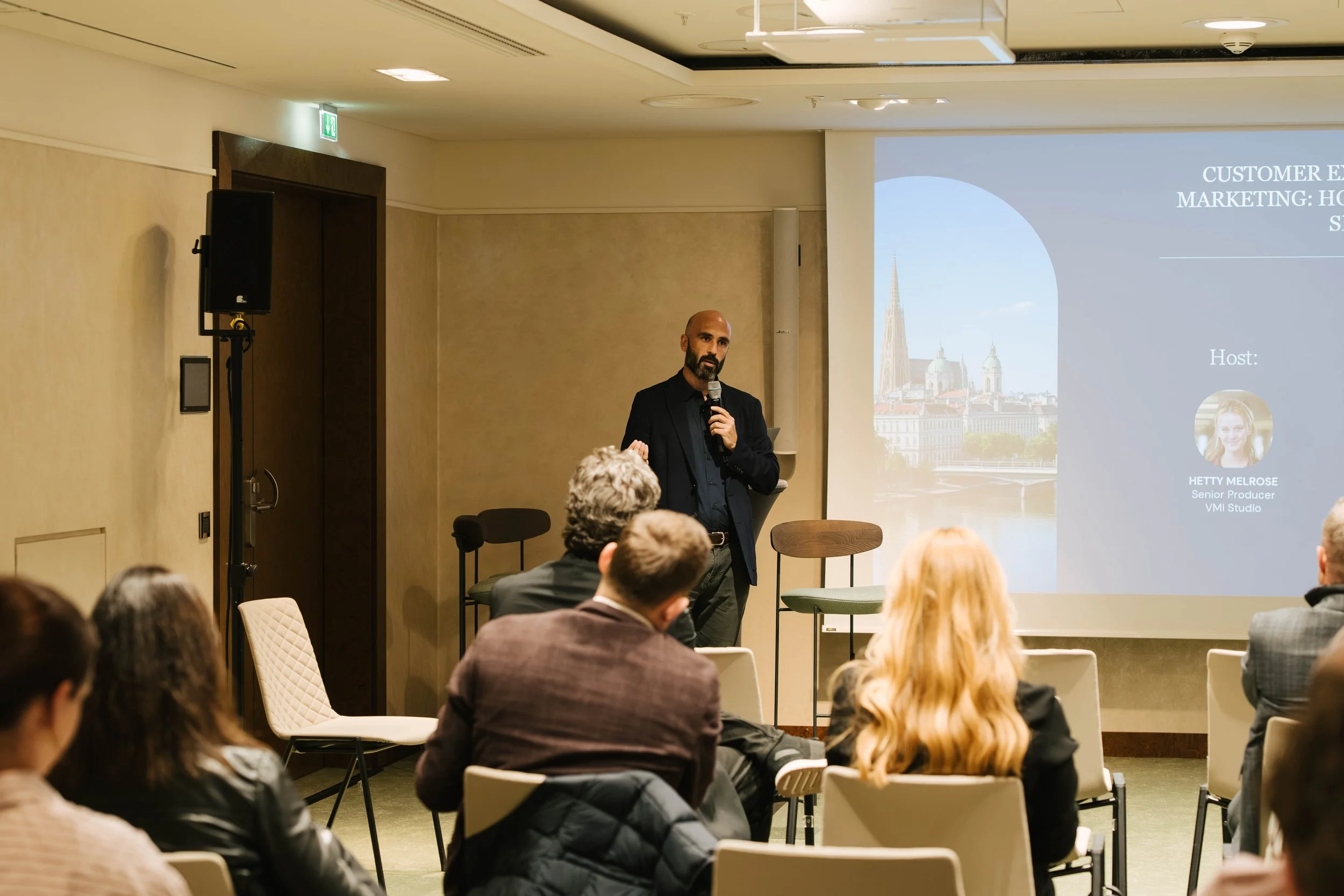 A man with a beard and bald head wearing a black blazer and shirt holds a microphone while presenting to an audience in a conference room. There is a projection screen displaying a presentation slide with a cityscape background and the name Hetty Mel