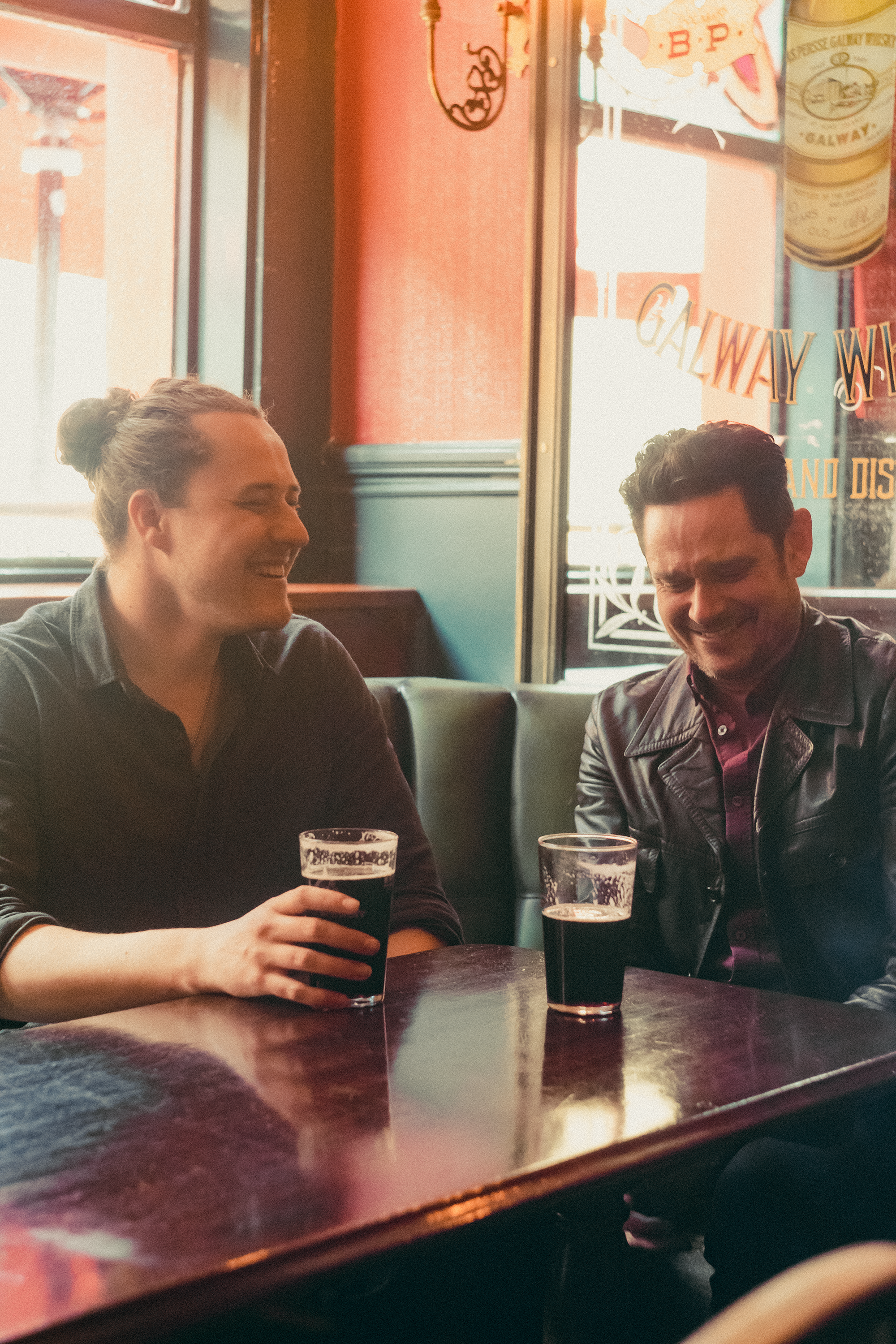 Two people sitting at a bar, holding glasses of dark beer, and smiling. They are inside a cozy pub with warm lighting and red walls. The woman on the left has her hair tied back in a bun, and the man on the right has dark, curly hair and is wearing a