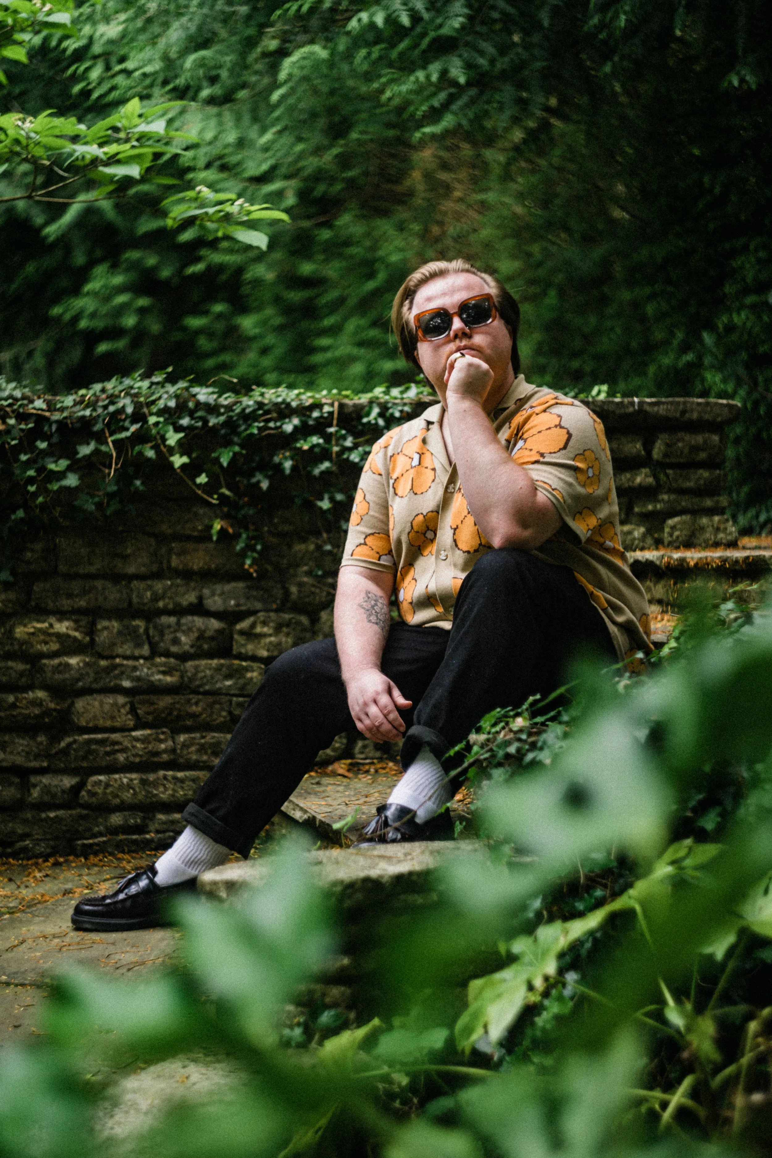 A person sitting on a stone ledge in a green outdoor setting, wearing large sunglasses, a beige shirt with orange floral patterns, black pants, white socks, and black shoes, with their hand resting on their chin and surrounded by lush green foliage.