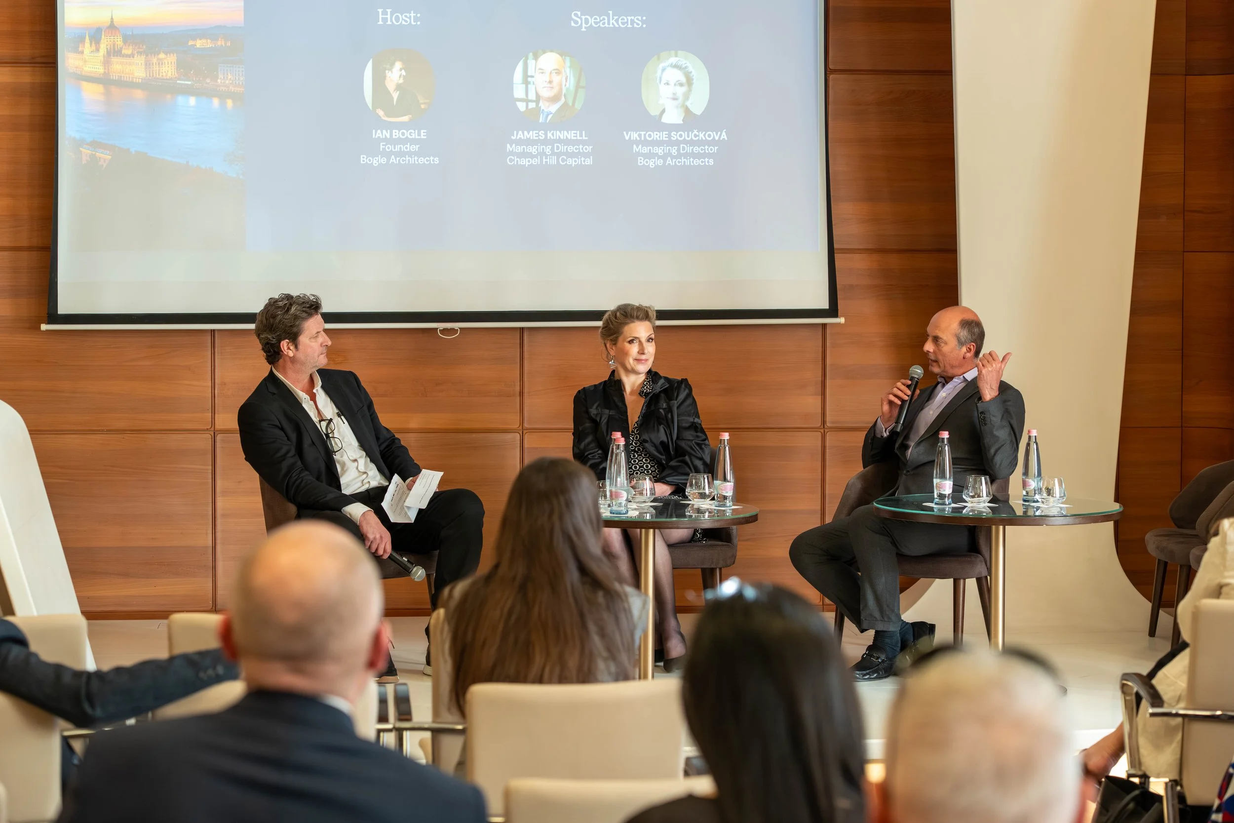 A panel discussion at a conference or seminar, with three speakers seated in front of a large screen displaying the event details. The audience is visible in the foreground.