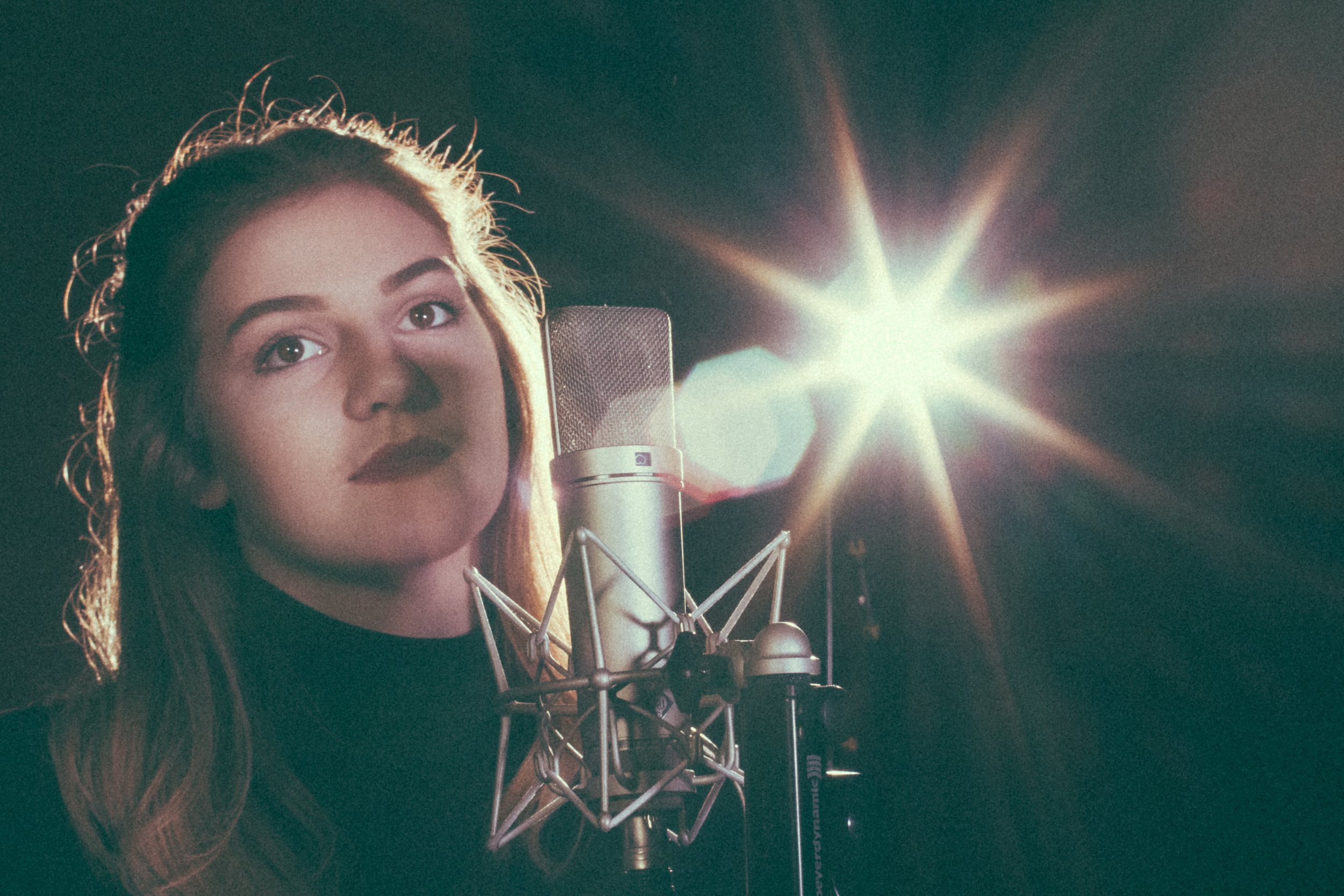 A young woman with red hair and wearing a black top is in a recording studio singing into a professional microphone.