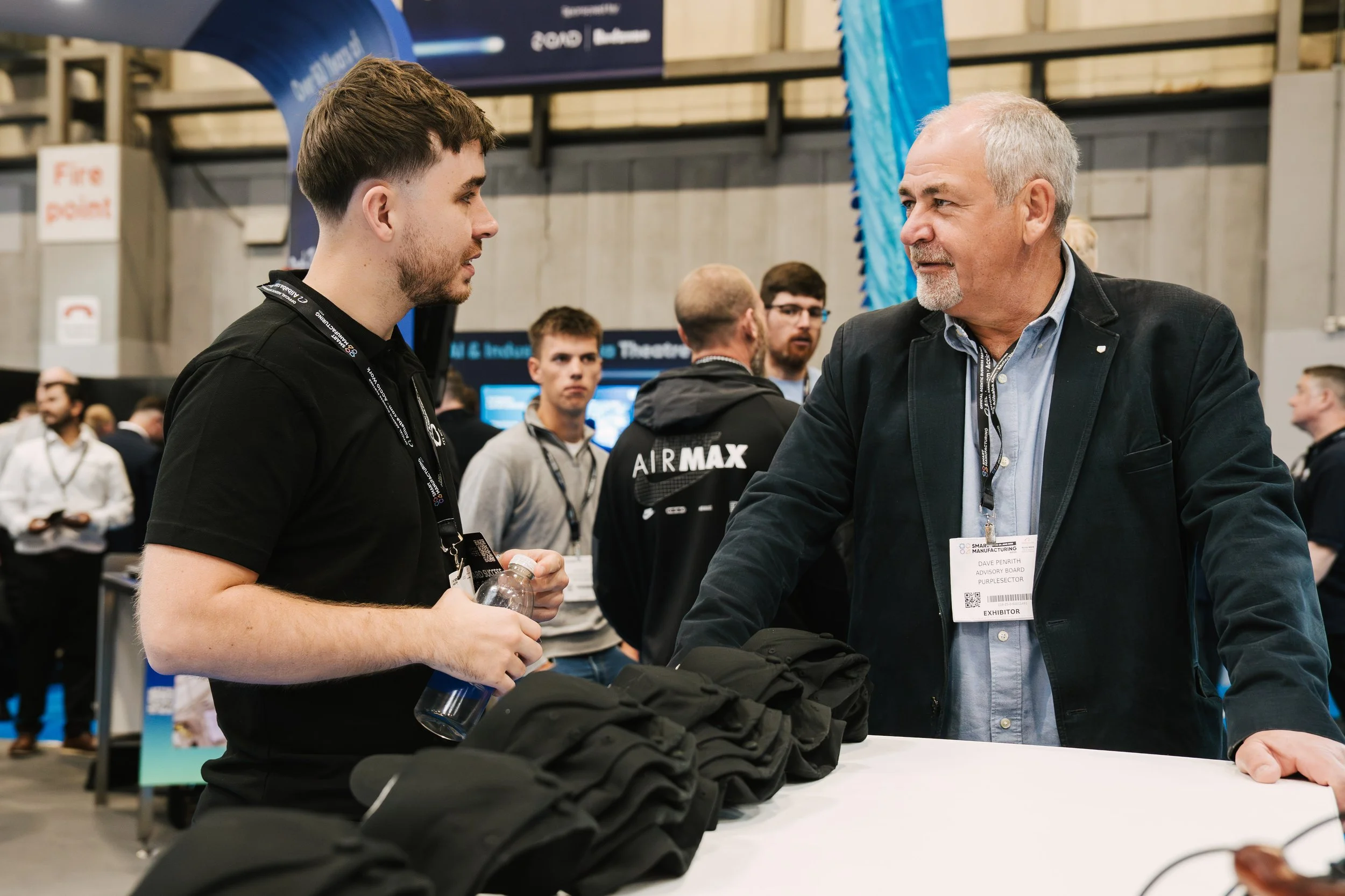 Two men are engaged in conversation at a professional event. One man, on the left, has short dark hair and a beard, wearing a black shirt and holding a water bottle. The other man, on the right, has short gray hair, a gray beard, and is wearing a dar