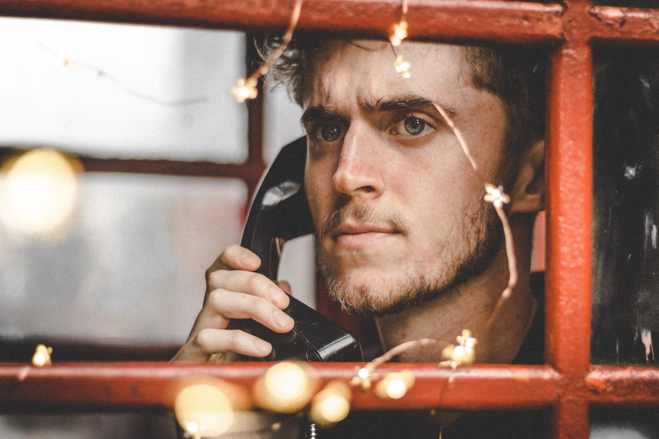 A young man with a beard and curly hair is looking through a red metal barrier while holding a black telephone receiver to his ear, with string lights decorating the foreground.