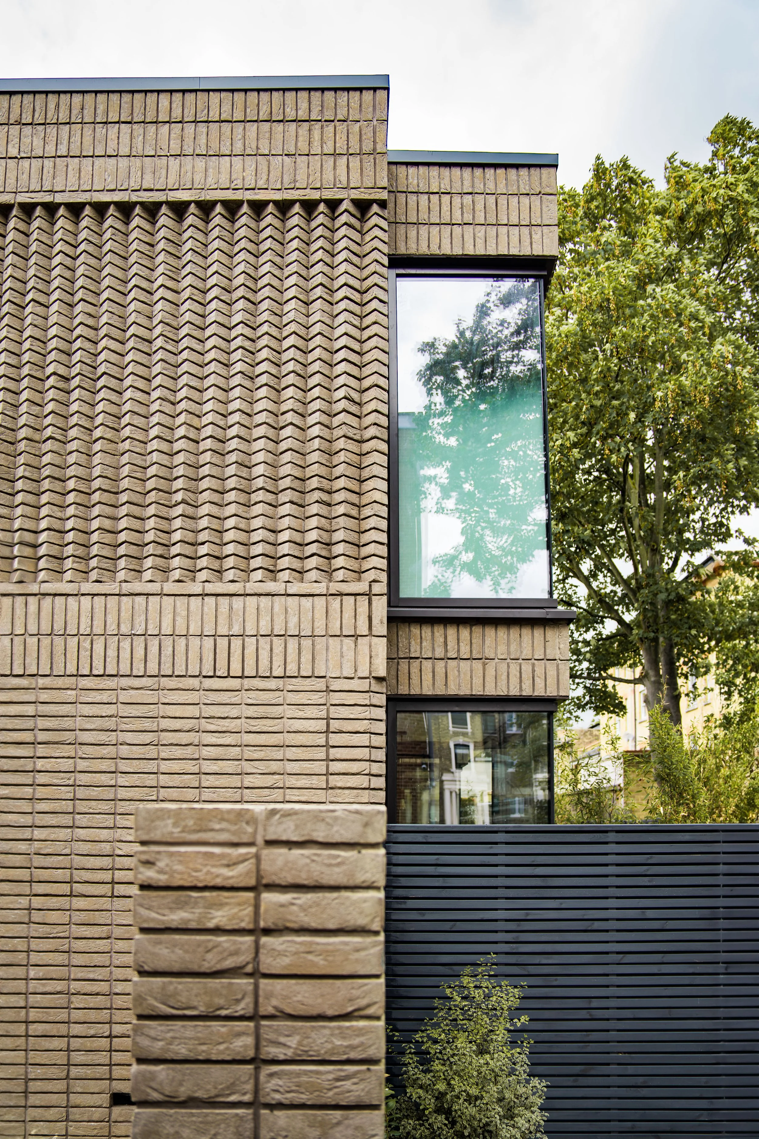 Close-up view of a modern building exterior with beige brick walls, large rectangular windows reflecting trees, and a black slatted panel, with greenery and trees in the background.