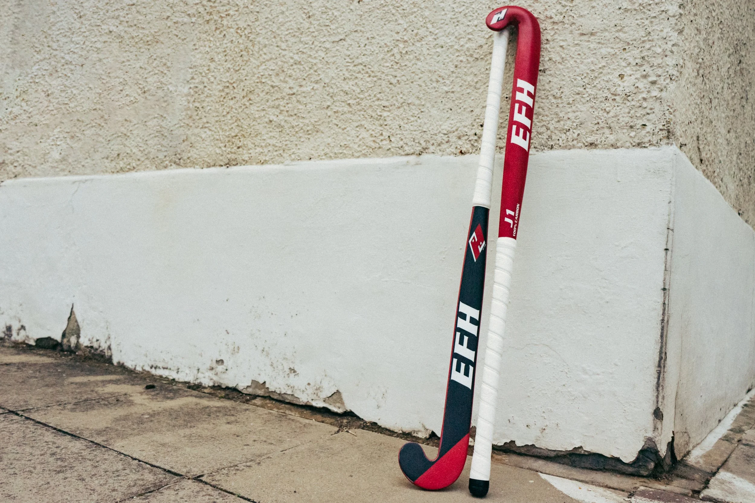 A Red and black hockey stick leaning against a white and beige wall with a concrete sidewalk in front.