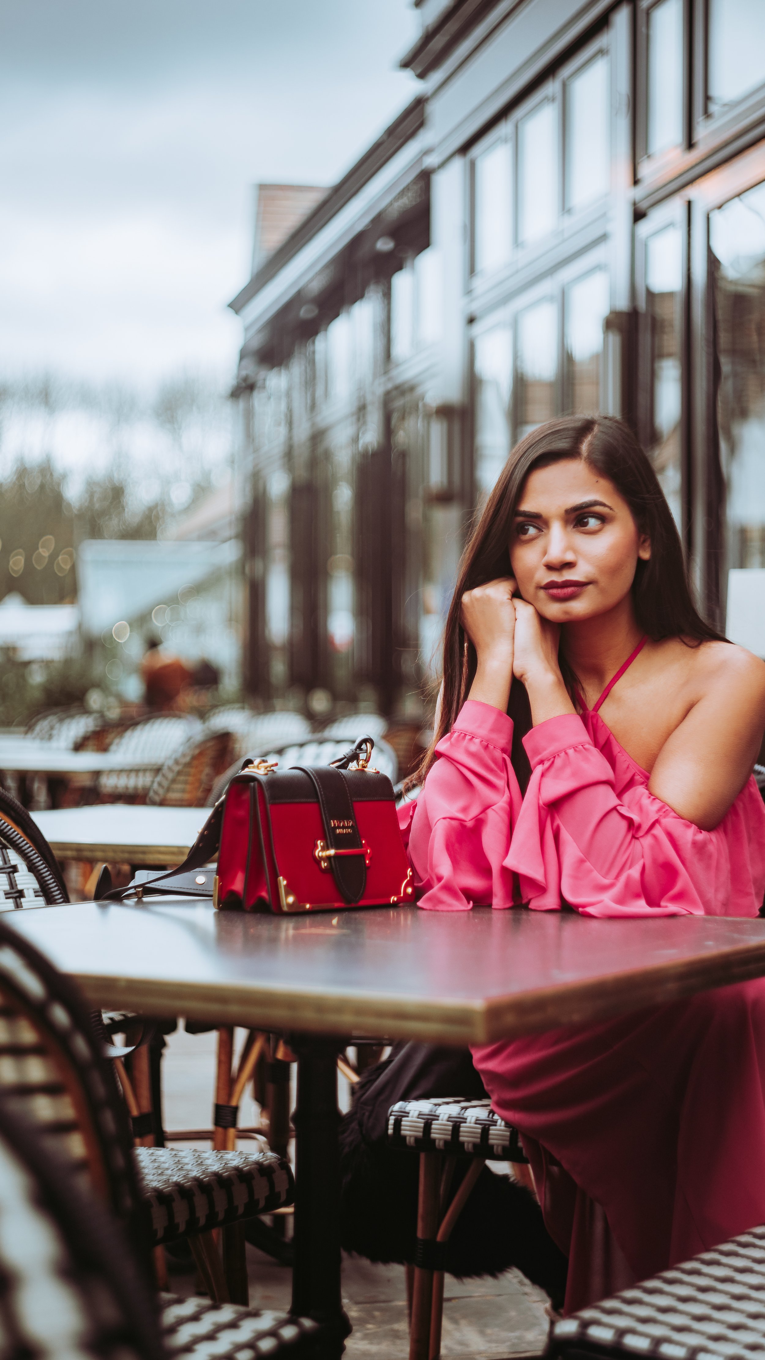 A woman in a pink dress sitting at an outdoor café table, resting her chin on her hands, with a red handbag on the table and a glass of water in front of her.
