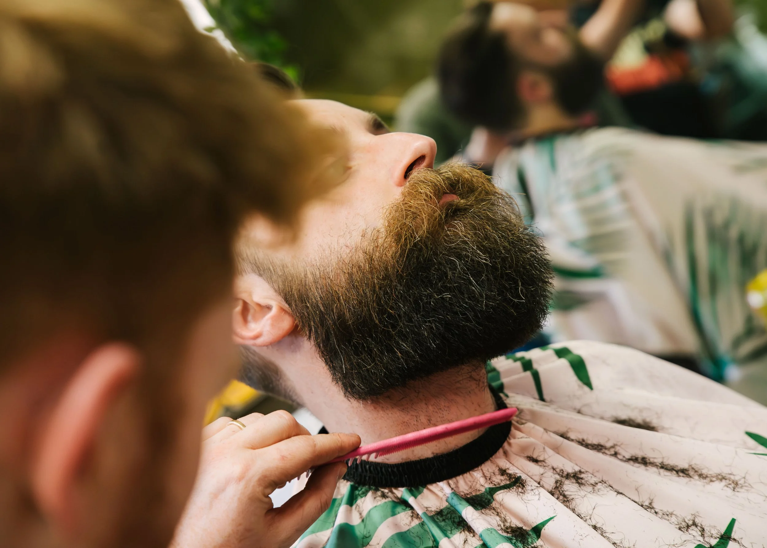 Close-up of a man's face and beard, with eyes closed and hand on chest, person sitting among others, wearing a patterned shirt with green and white colors, in a group setting.
