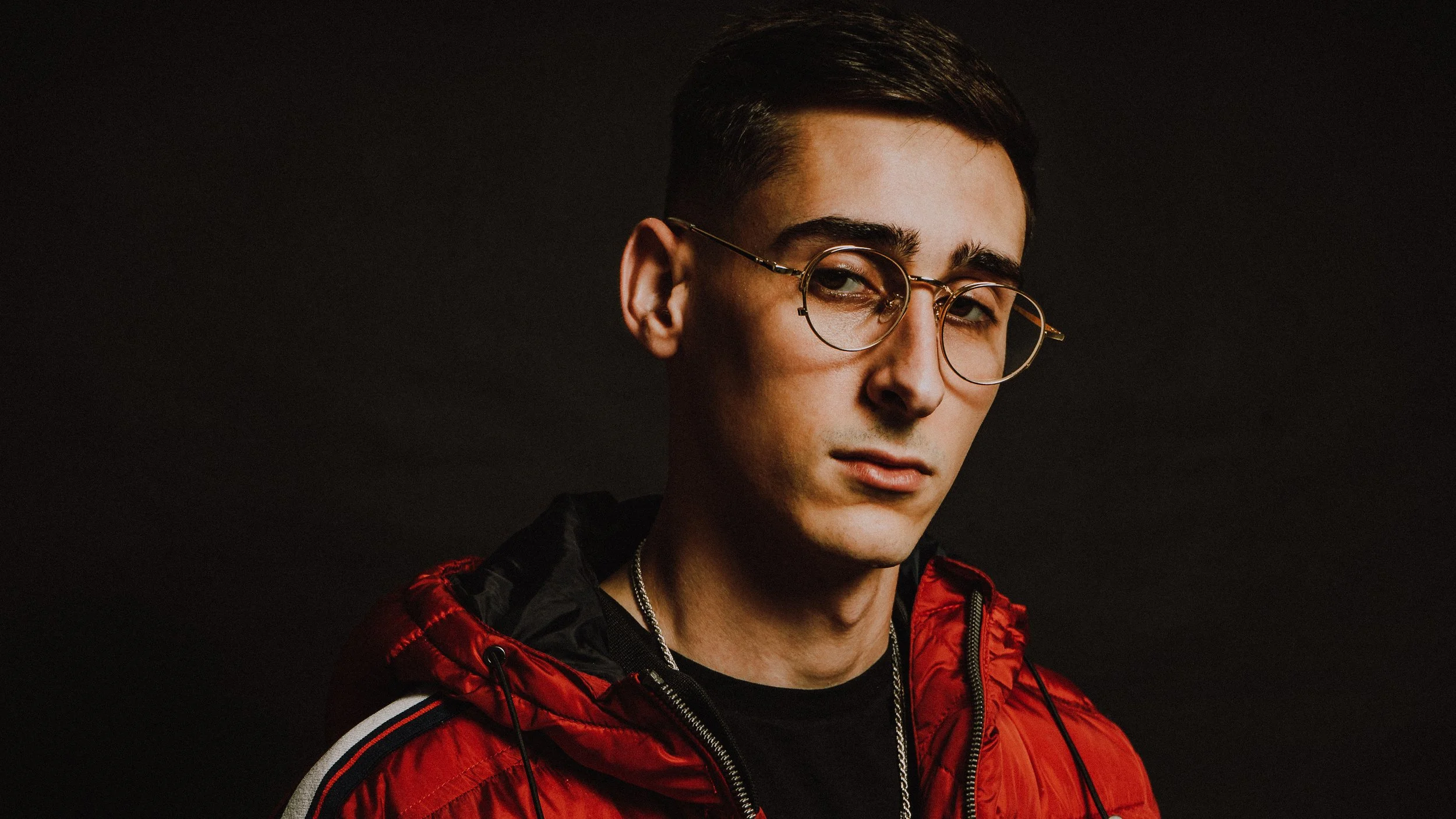 A young man with dark hair, wearing glasses, a black shirt, and a red jacket, looks to the camera with a serious expression. Dark background.