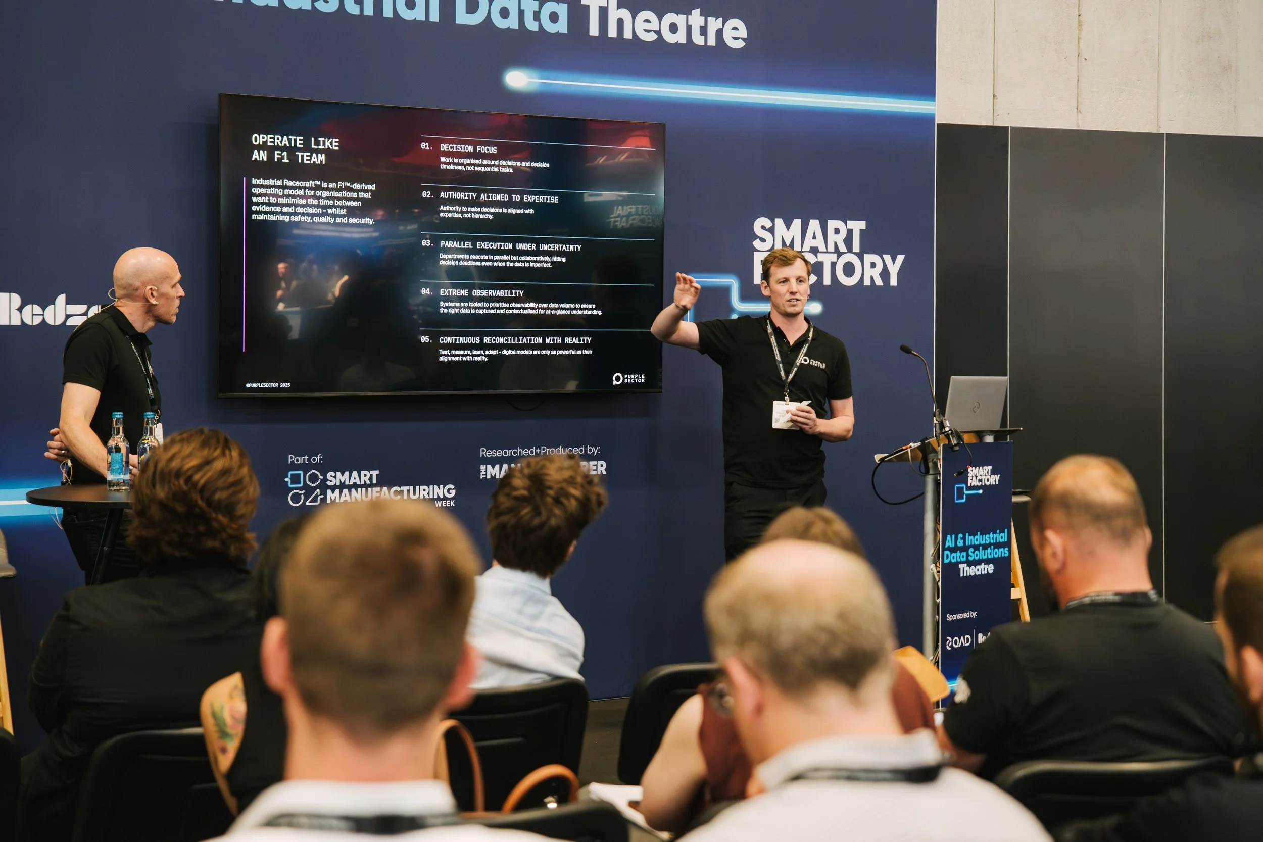 A man standing at a podium giving a presentation at a conference, with a large screen behind him displaying slides about AI and industrial data solutions, and an audience seated in front of him.