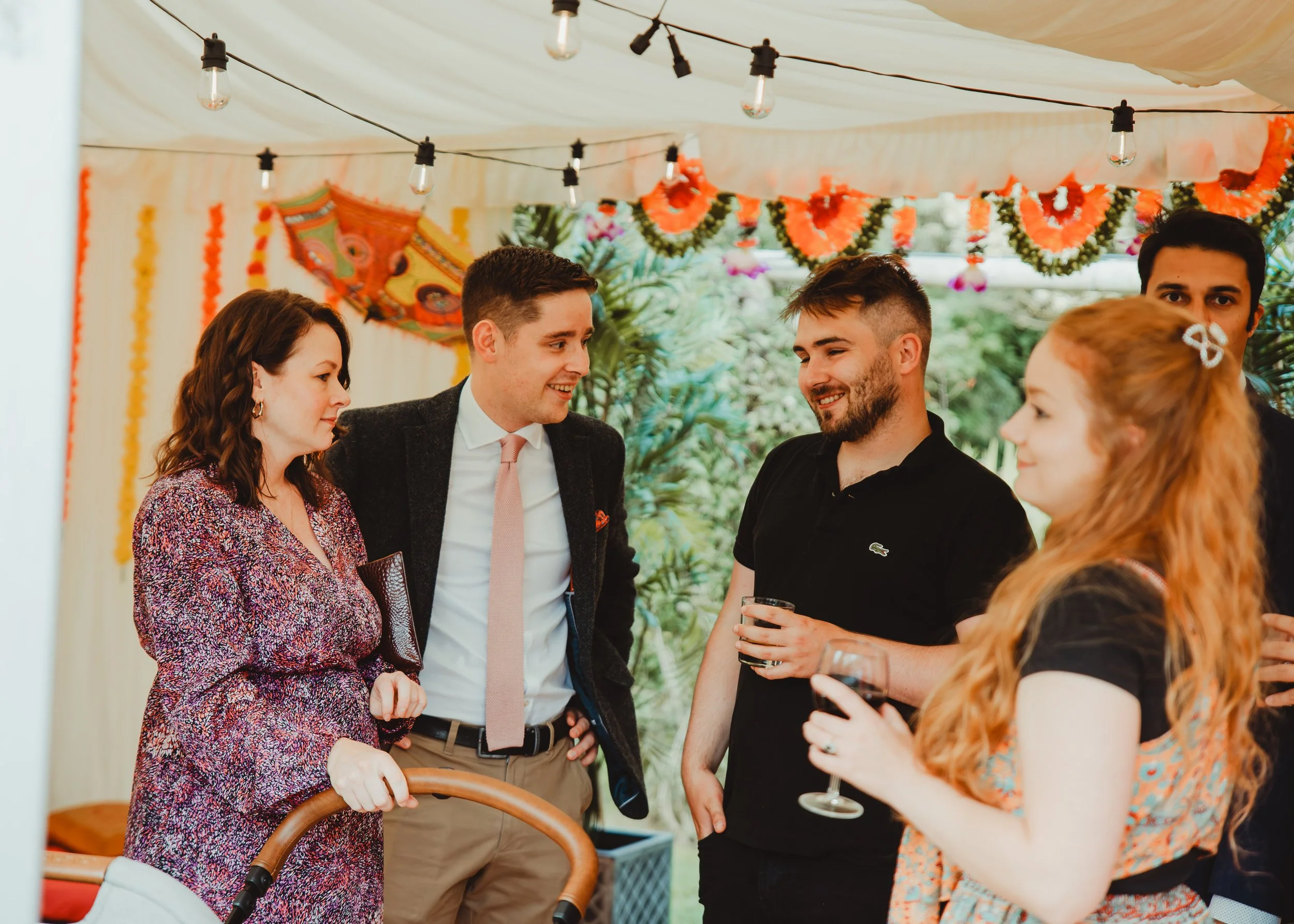 A group of five adults at a social gathering under a decorated outdoor tent, having conversations. There are colorful garlands and string lights overhead. One woman is holding a drink, and a man in a black shirt has a Lacoste logo.