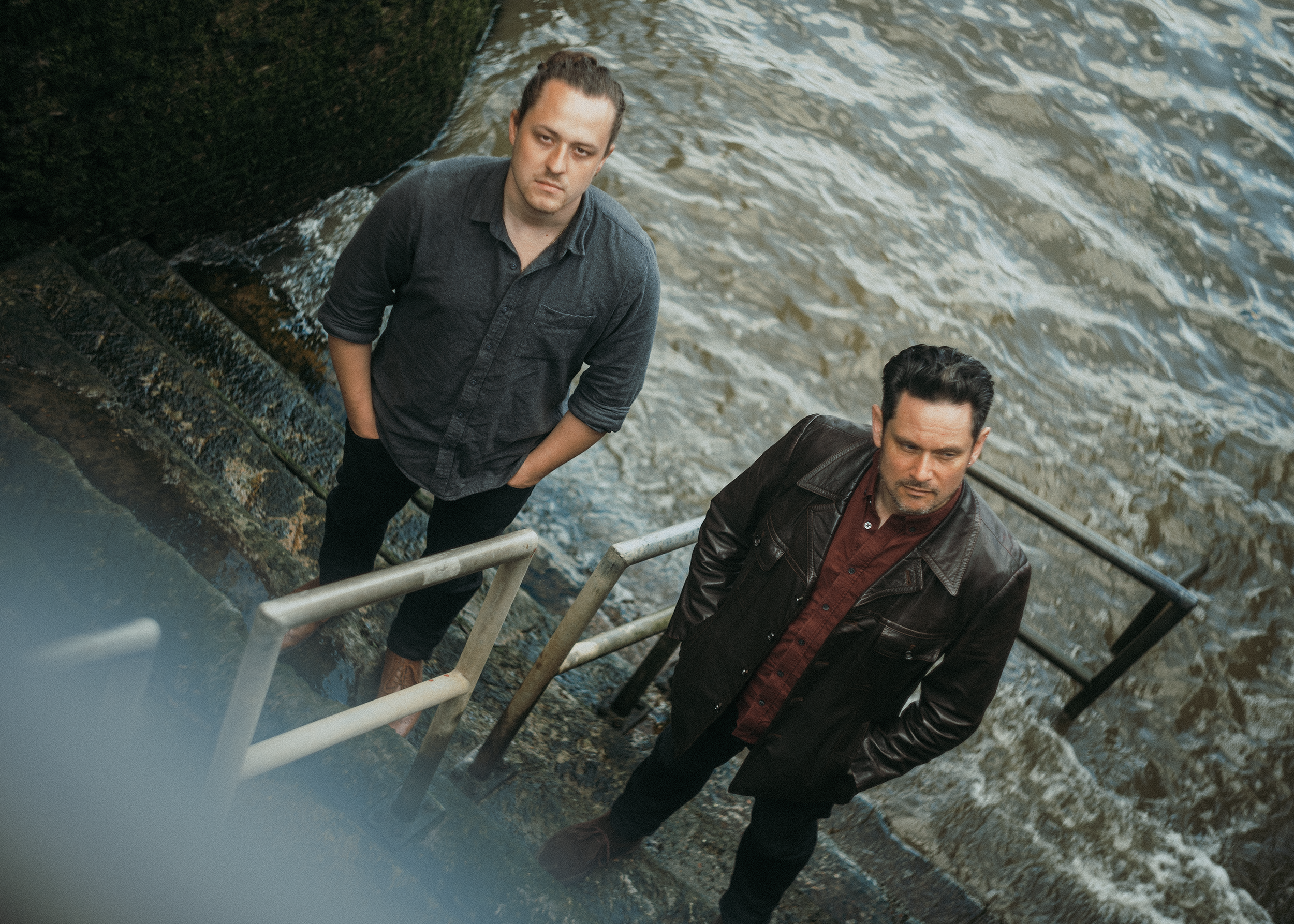 Two men standing on a staircase near water, looking up at the camera, with a dark wall or structure on the left side.