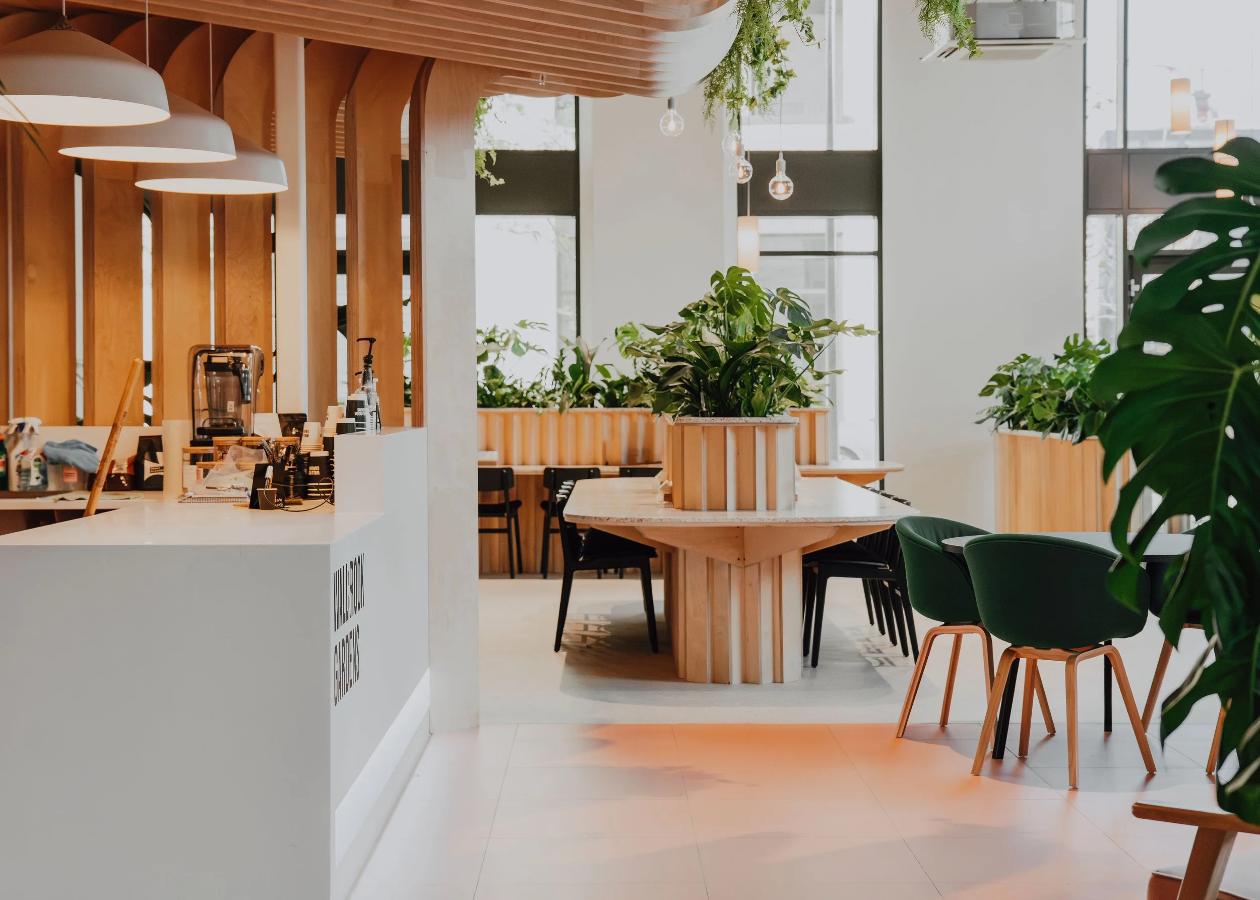Modern interior cafe with wooden accents, large windows, and lush green plants, including hanging plants and potted plants on tables, with pendant lights and seating areas.