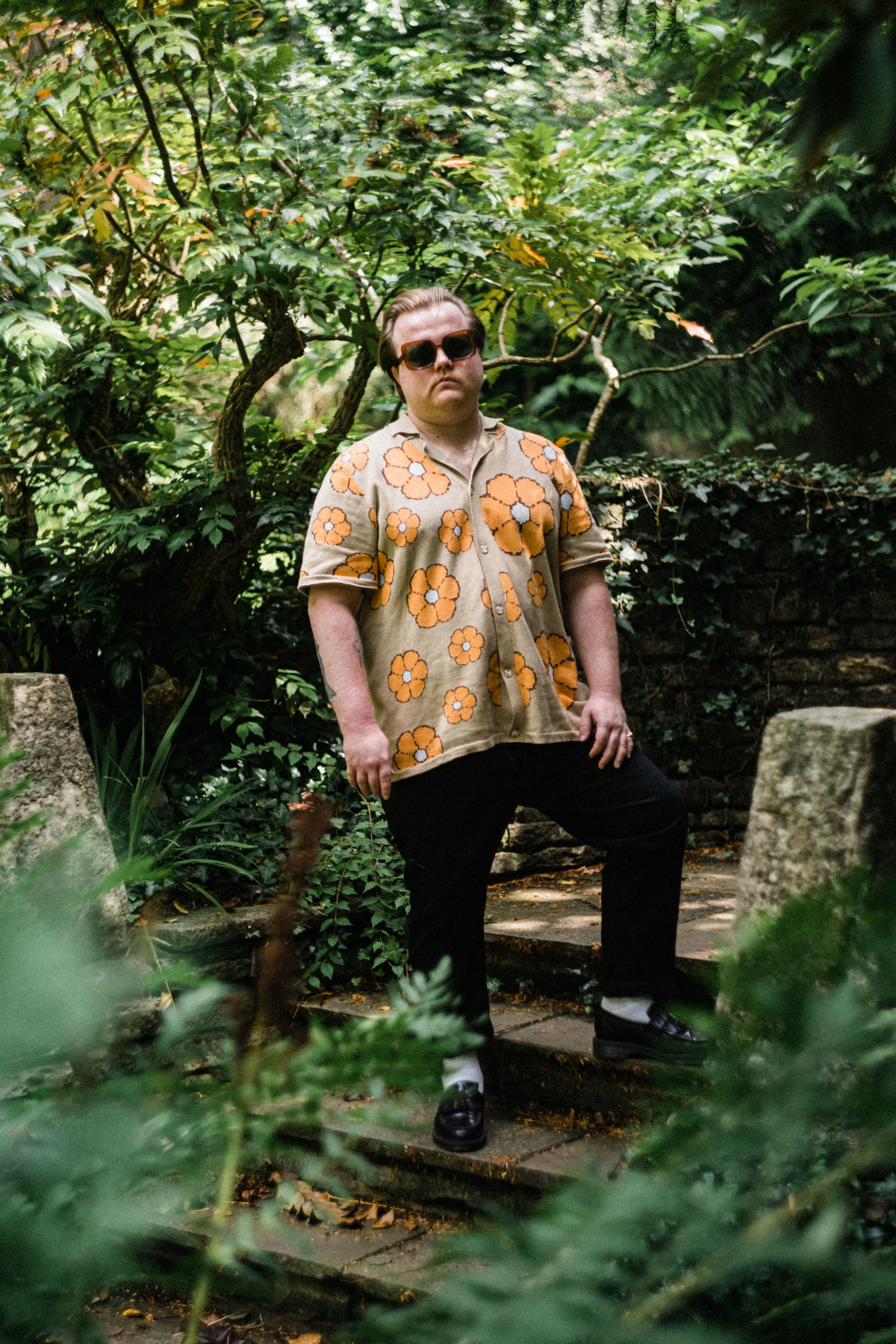 A man standing in a lush garden surrounded by trees and green foliage, wearing sunglasses and a beige shirt with orange flowers, black pants, and black shoes.