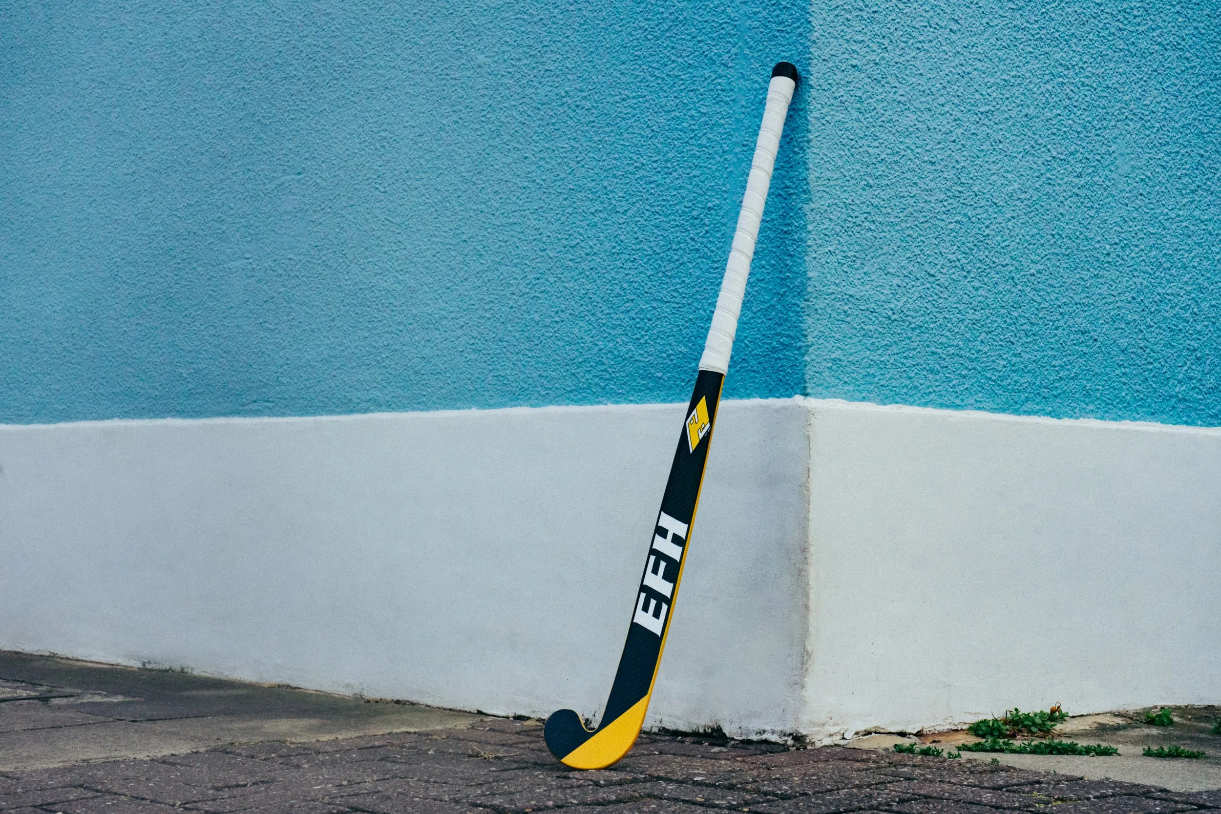 Hockey stick leaning against a corner of a blue textured wall with a white baseboard. The stick is black and yellow with the words "EHF" visible.