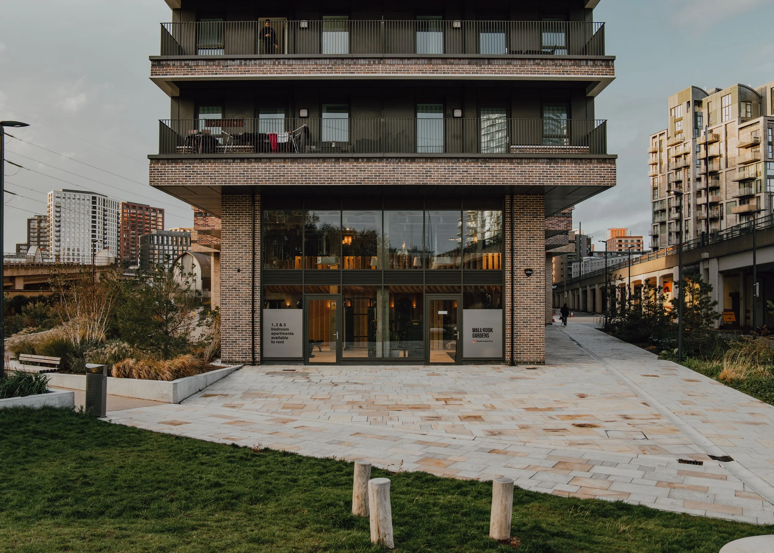 Modern brick building with large glass windows and balconies, surrounded by a paved walkway and landscaped greenery.
