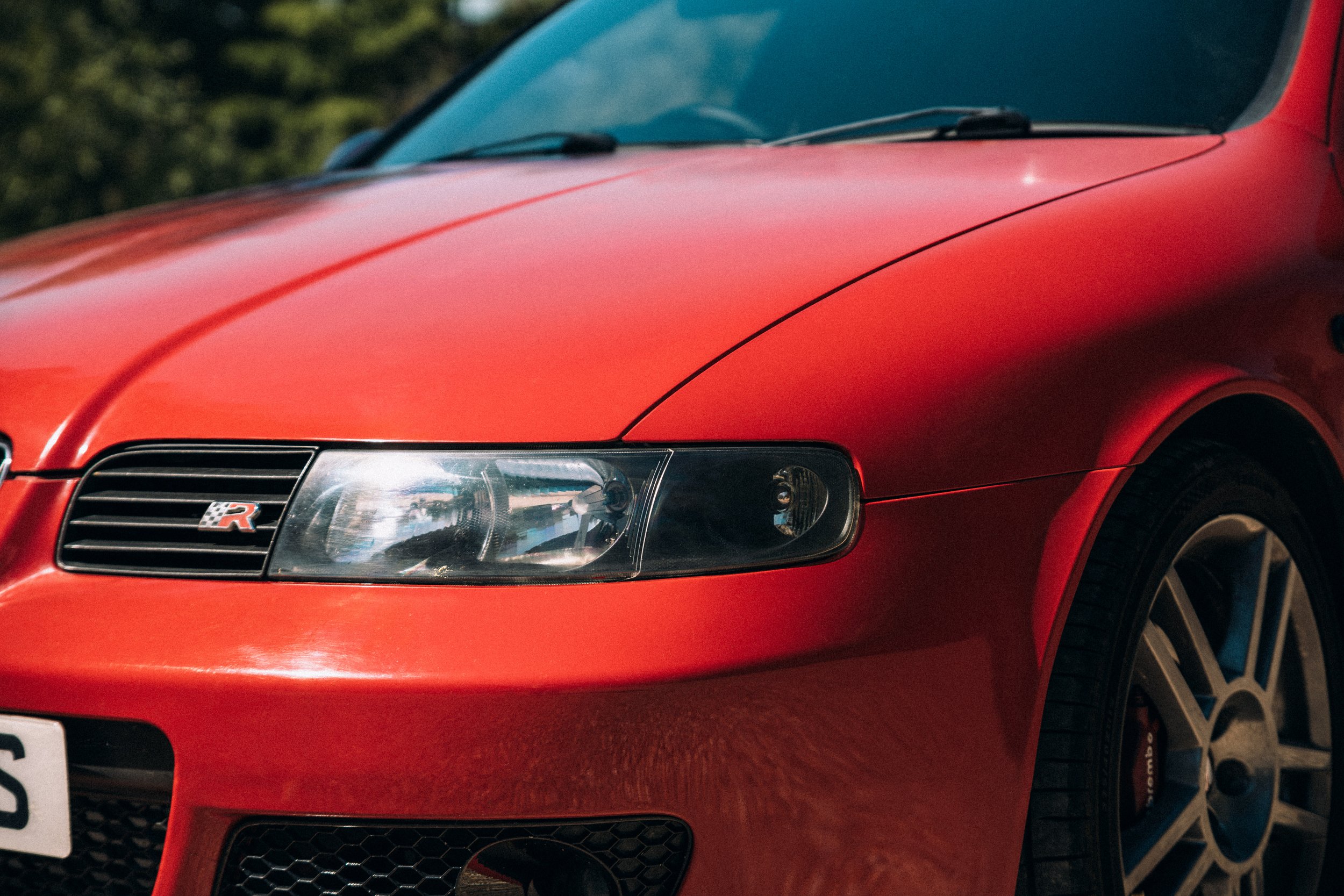 Close-up of the front of a red sports car with an R badge, headlight, and tire visible, parked outdoors with trees in the background.