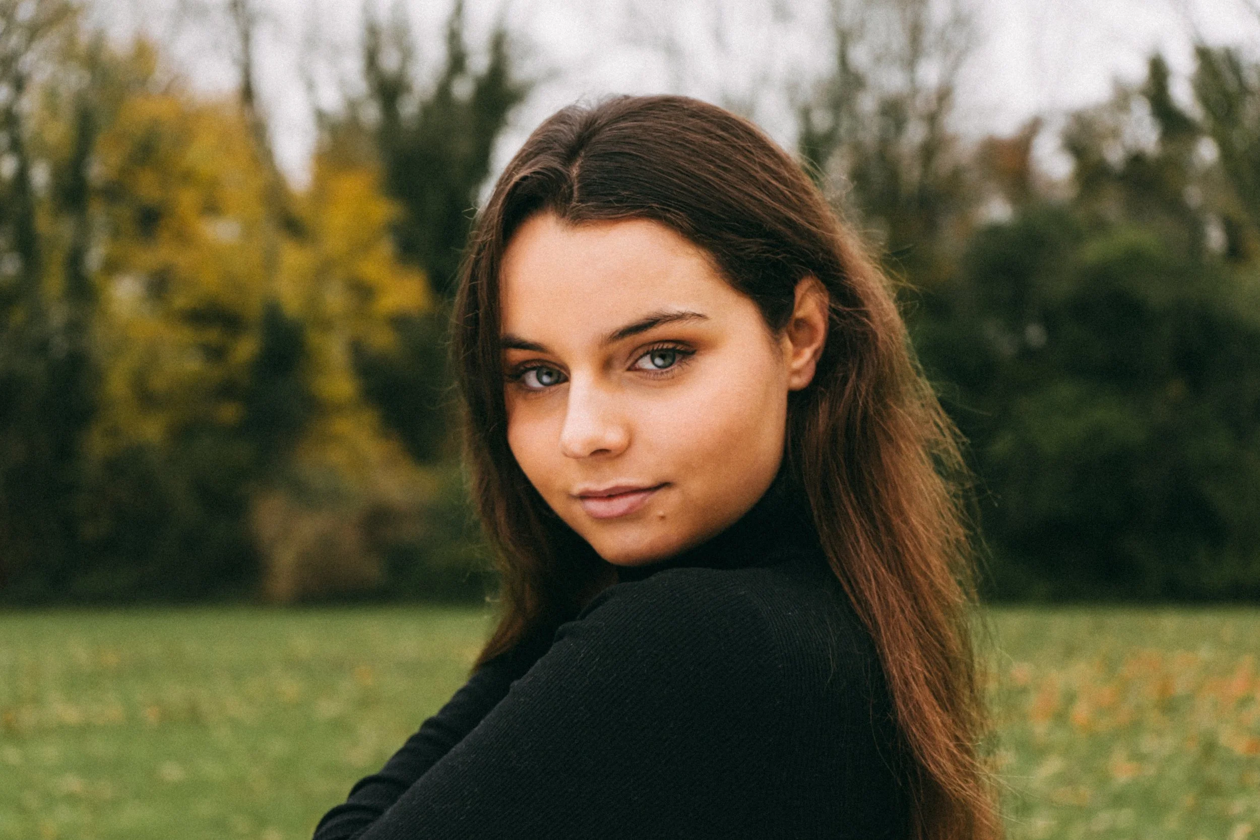 A young woman with brown hair and blue eyes posing outdoors in a park during autumn, wearing a black top.