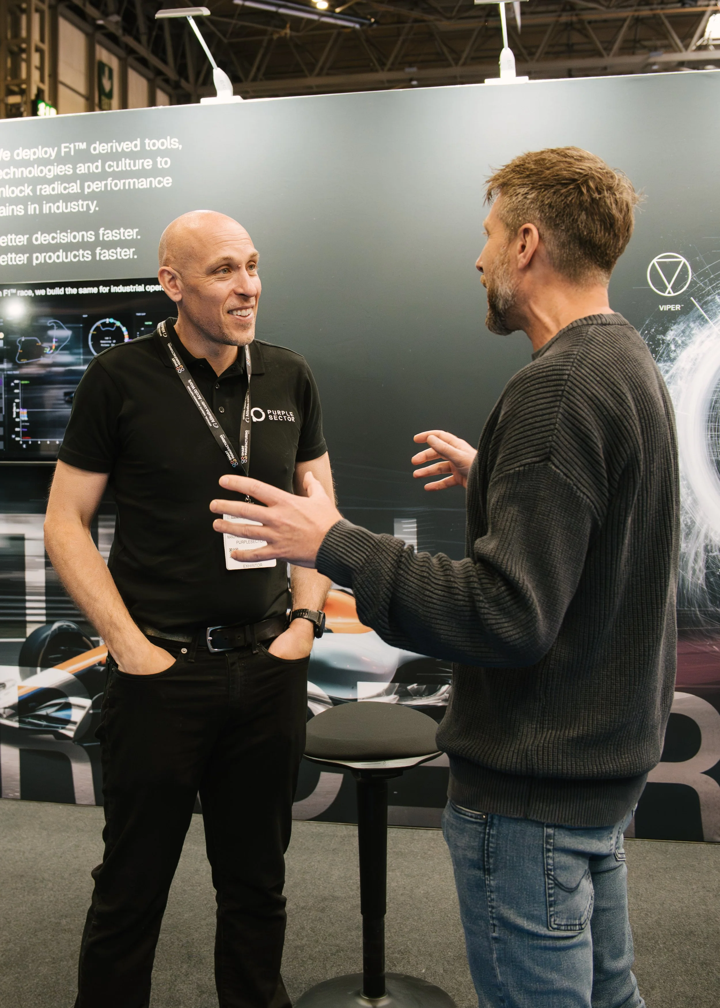 Two men are engaged in a conversation at an exhibition booth; one man is wearing a black shirt with the logo 'Purple Sector' and a lanyard, while the other man is dressed in a dark sweater and jeans. Behind them is a large display panel with partiall