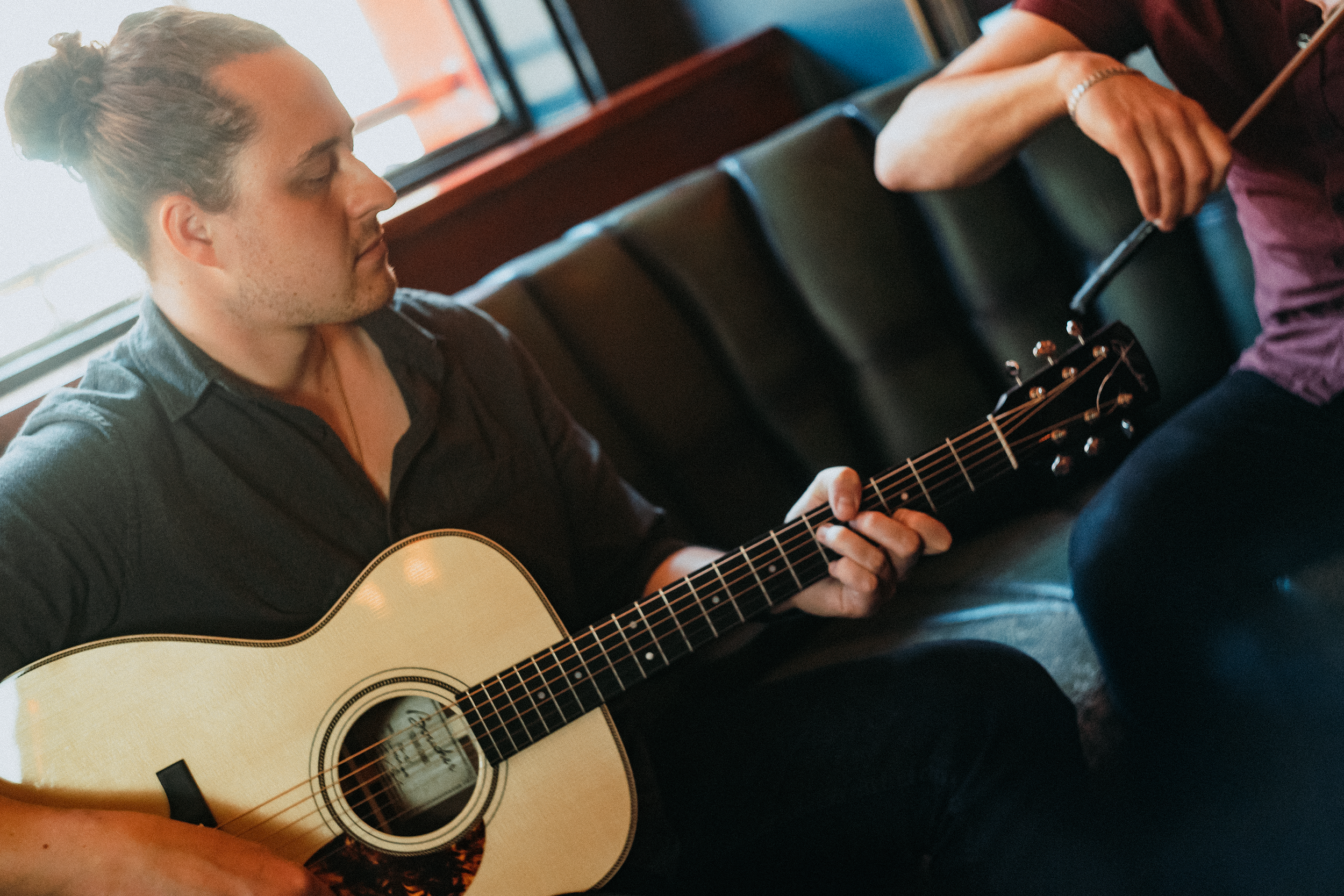 A young man with long hair tied in a bun playing an acoustic guitar in a cozy indoor setting.