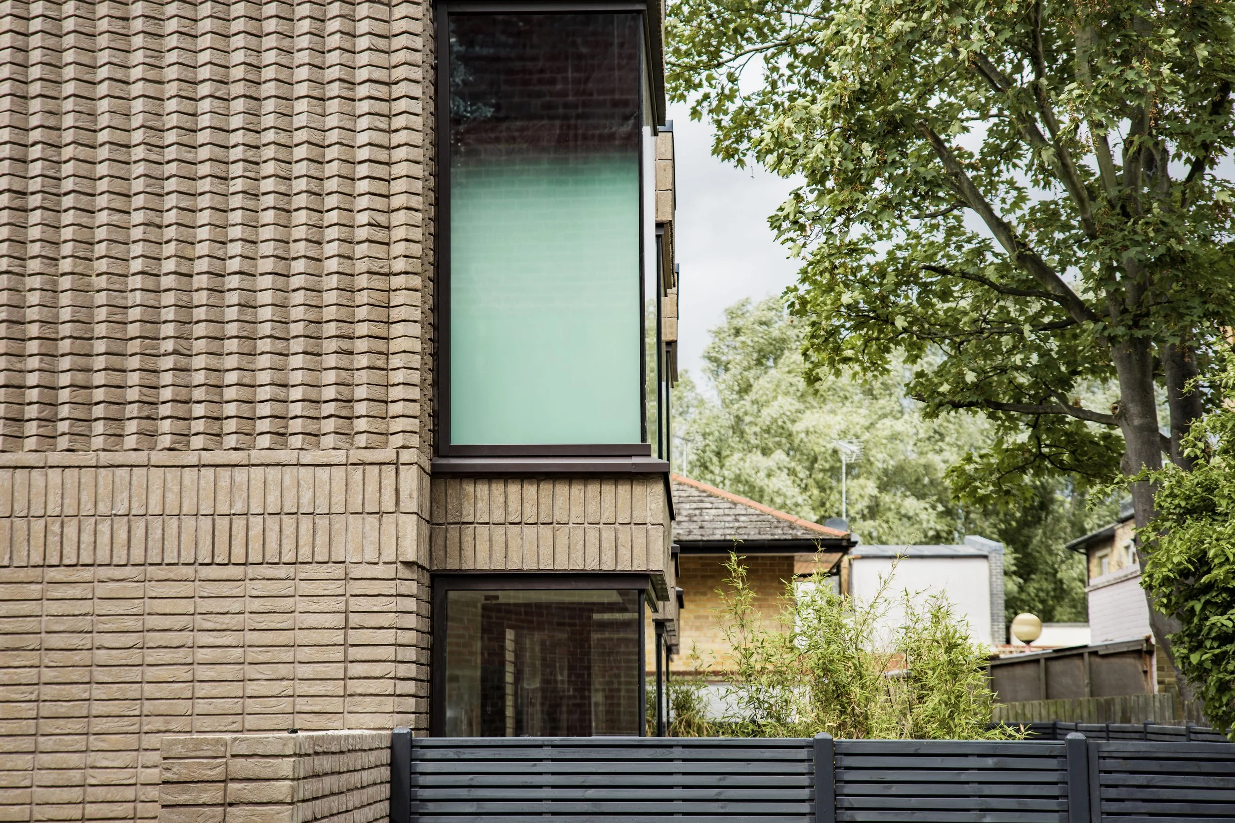 Close-up view of a modern house exterior with beige brick walls, large glass windows, and a balcony, surrounded by green trees and vegetation.