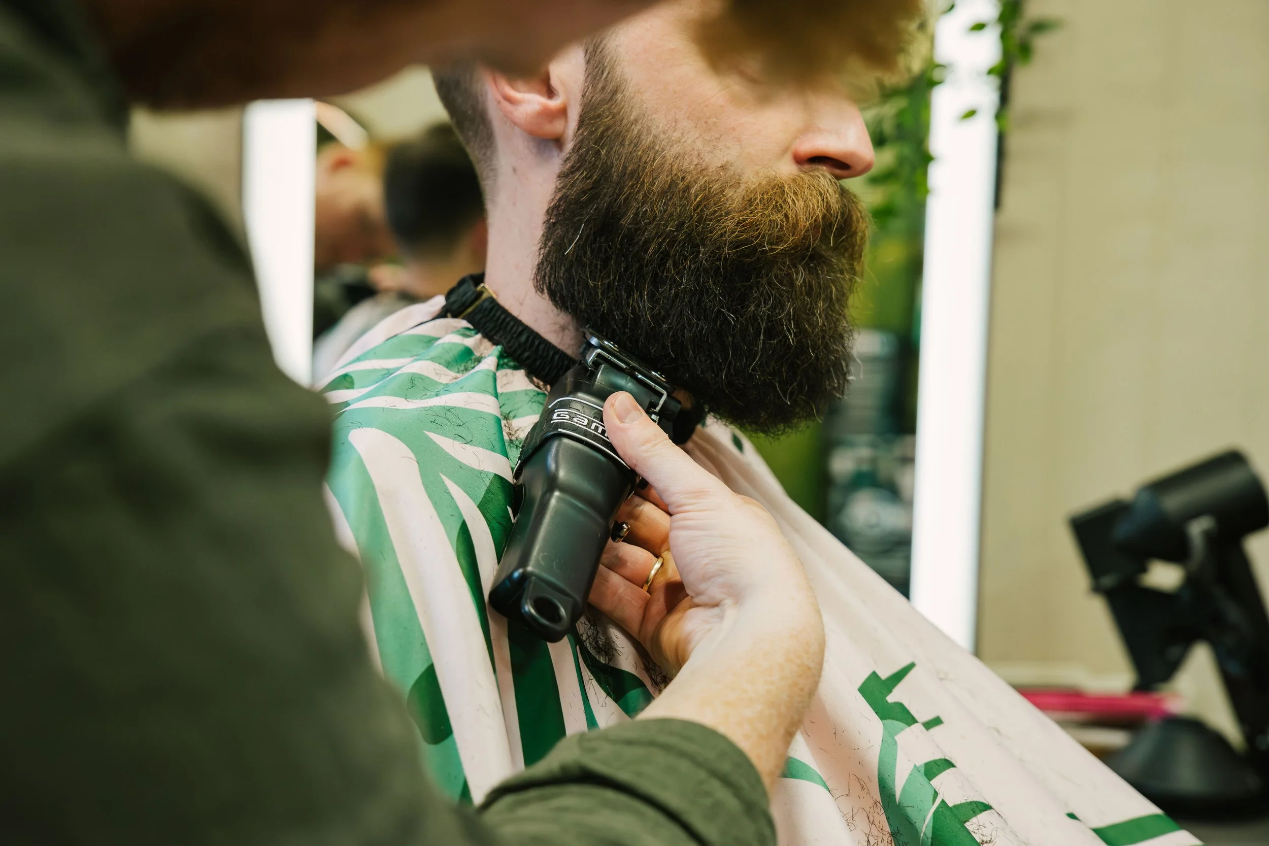 A man with a full beard is getting a haircut at a salon, with a barber trimming his beard.