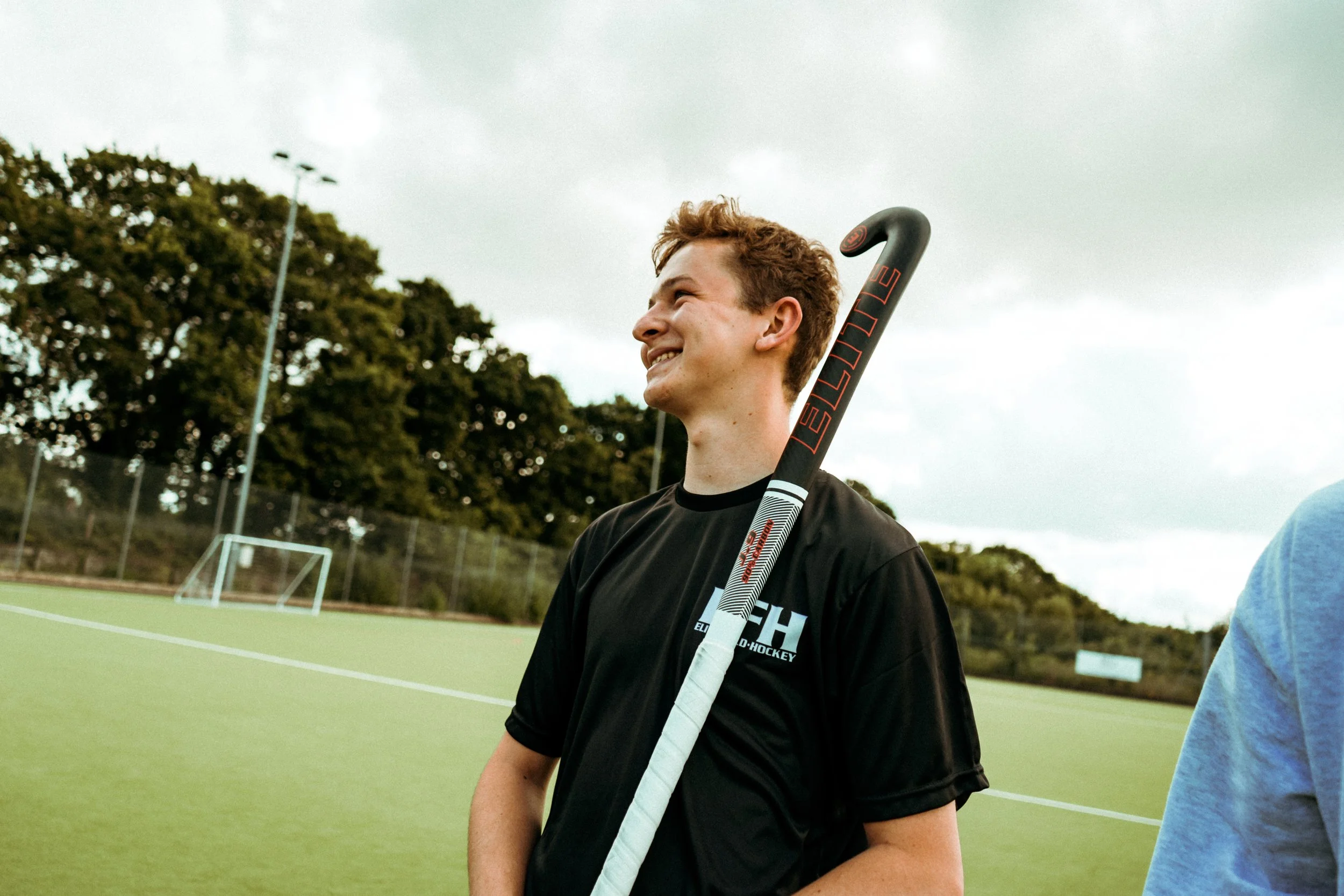 A young man with brown hair smiles on an outdoor hockey field, holding a field hockey stick, with trees and a cloudy sky in the background.