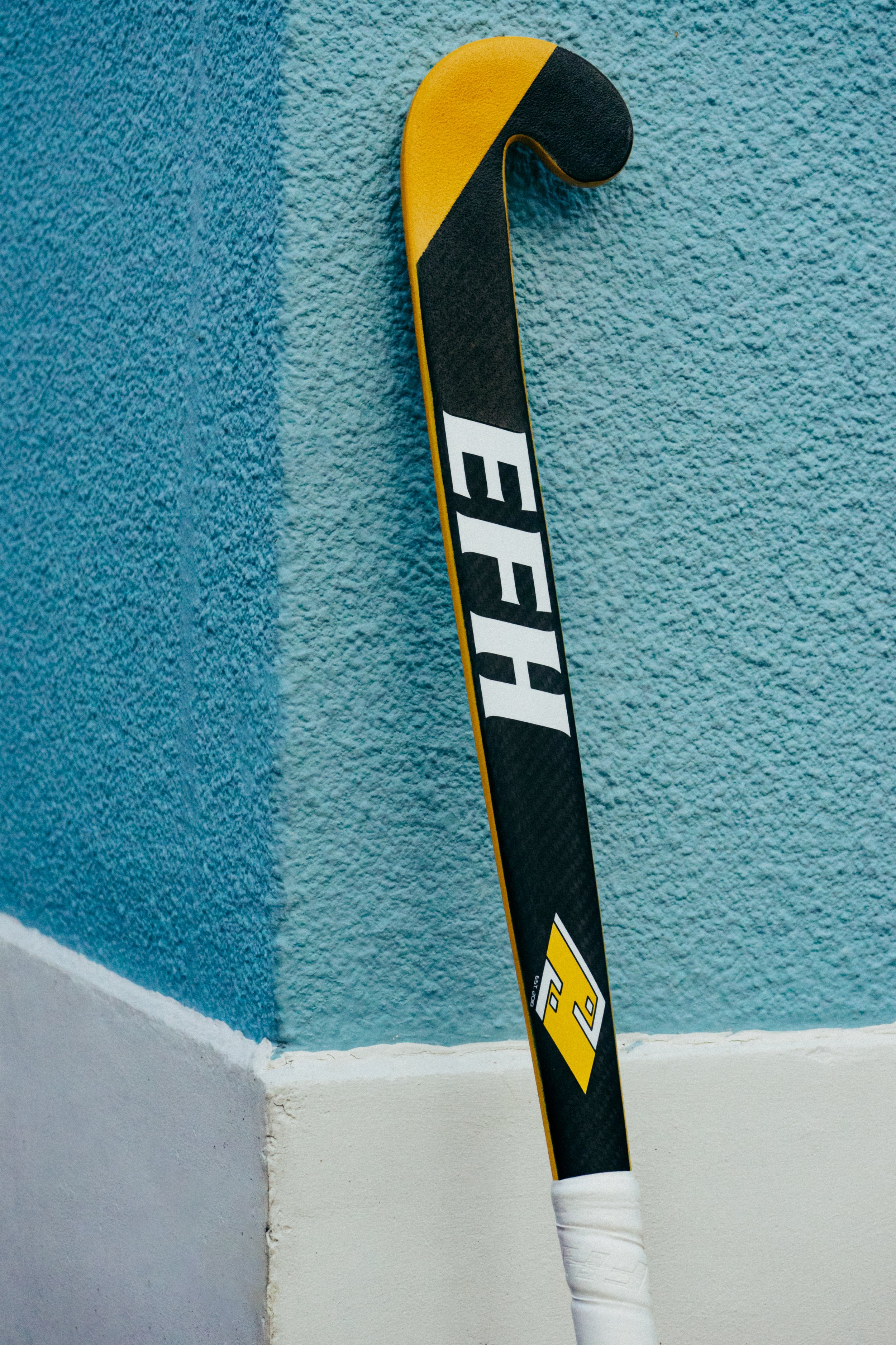 A hockey stick leaning against a textured wall painted with blue and teal. The stick has a black, yellow, and white design with the brand name partially visible.