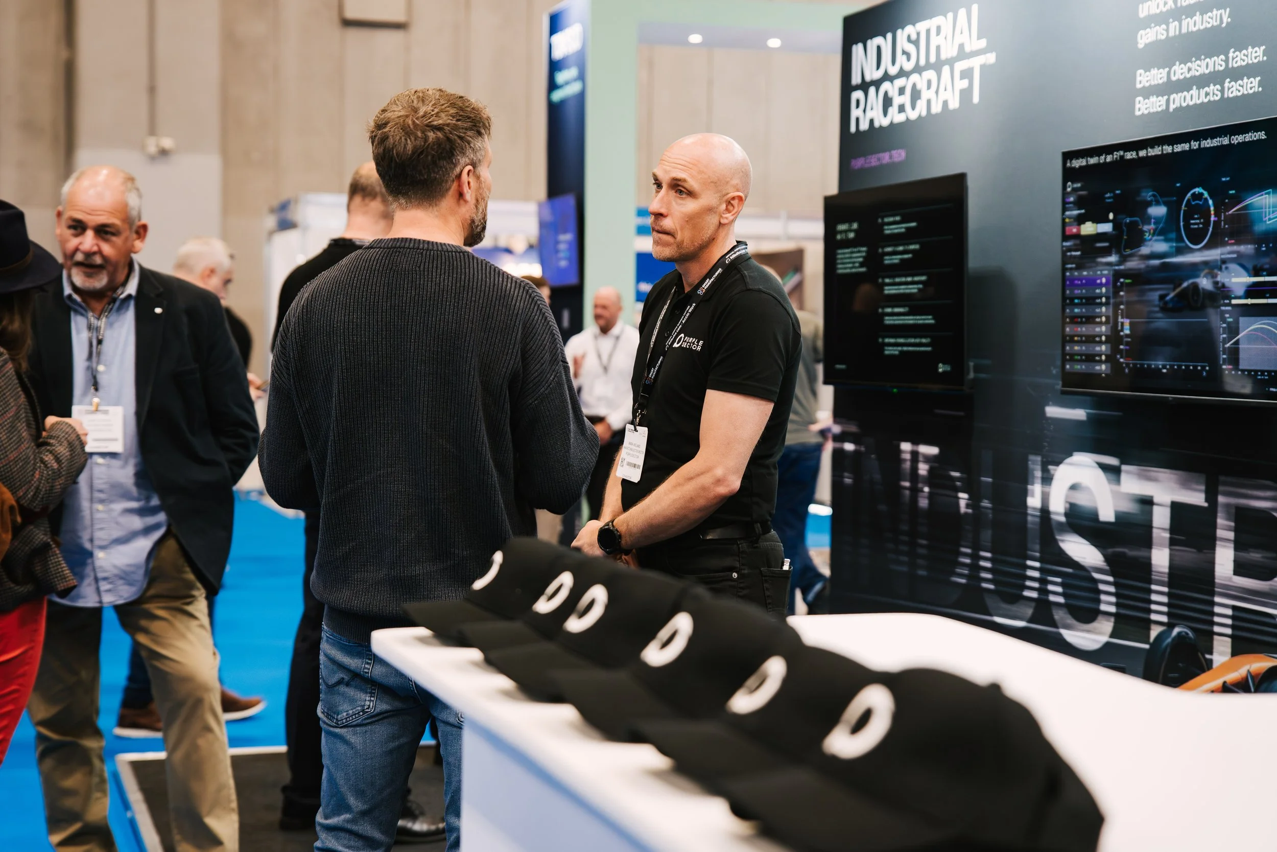 Two men are engaged in conversation at a trade show booth with the sign 'Industrial Racecraft' behind them. Other people are visible in the background.