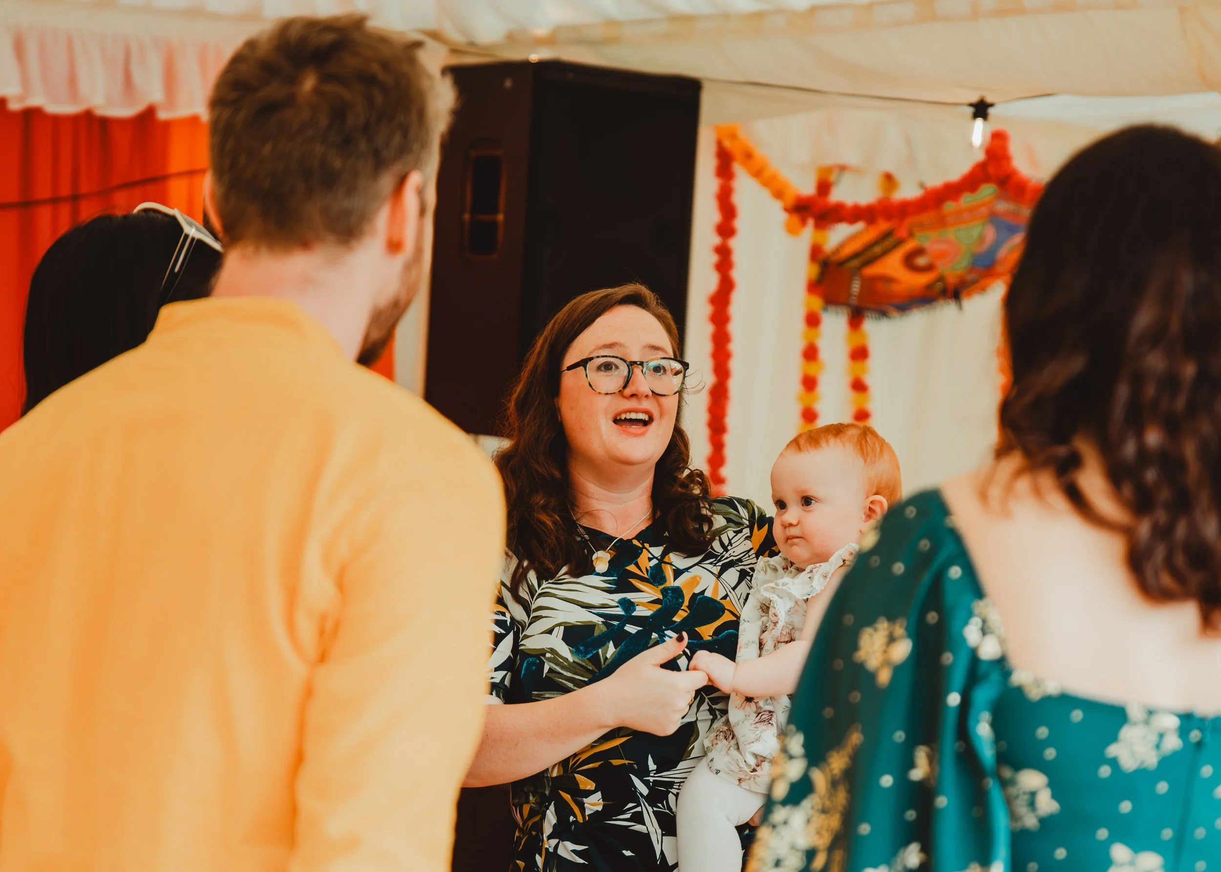 Group of people, including a woman holding a baby, celebrating at a festive event with orange and green decorations and a Chinese dragon puppet.