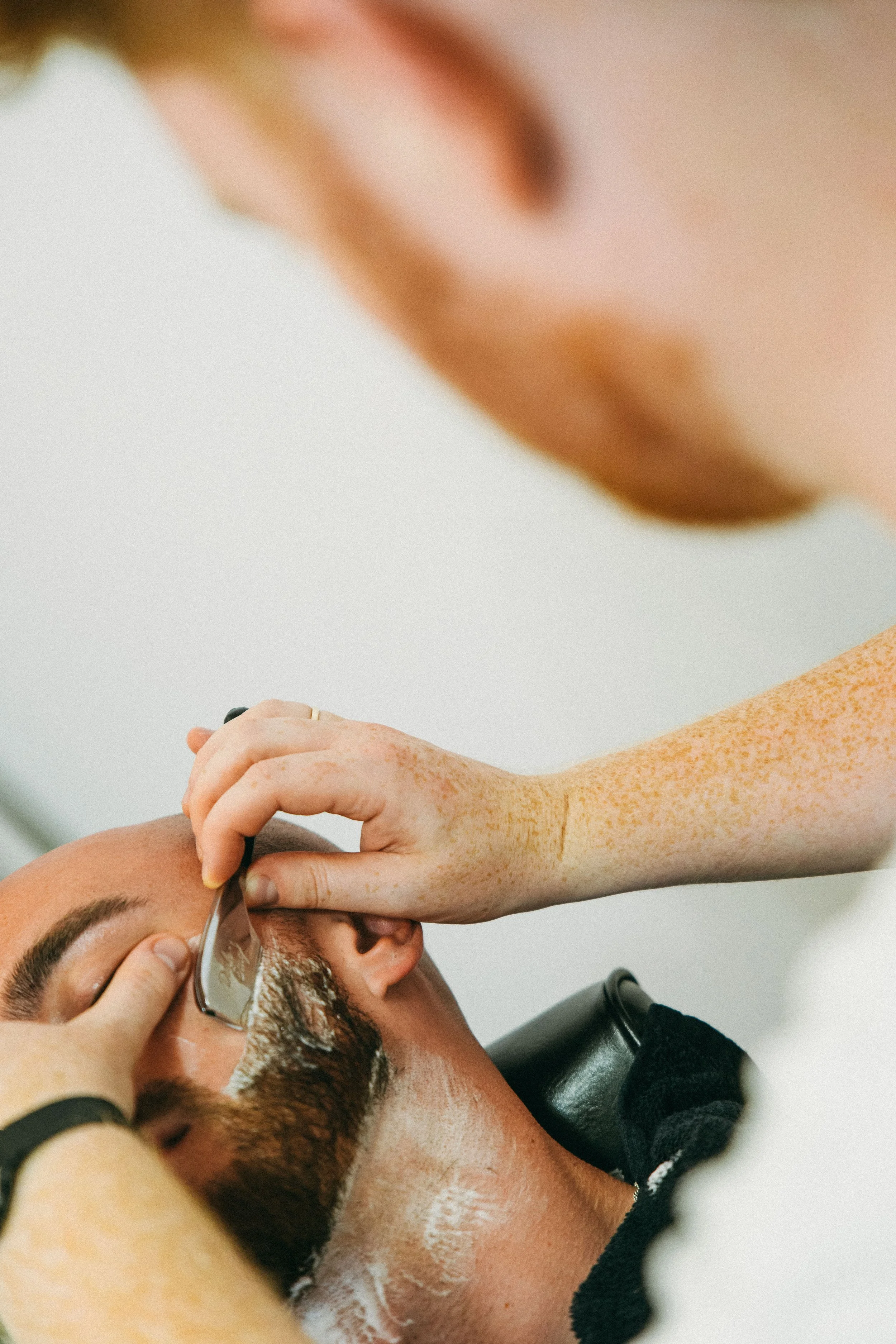 A person receiving a haircut or grooming at a salon, with a stylist working on their hair while they lie back with eyes closed.