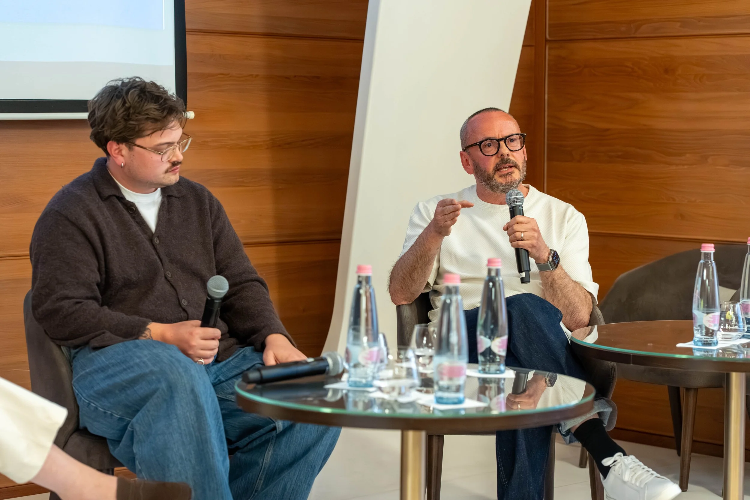 Two men seated at a panel discussion table. The man on the right is speaking into a microphone, gesturing with his hand. The man on the left is looking down, holding a microphone. The table has bottles of water. The room has wood-paneled walls and a 