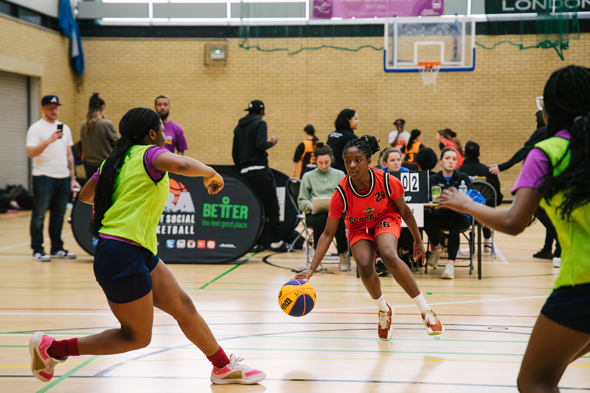 Young female basketball player in red uniform dribbling a basketball during a game in an indoor gym, flanked by two players in neon yellow and purple uniforms.
