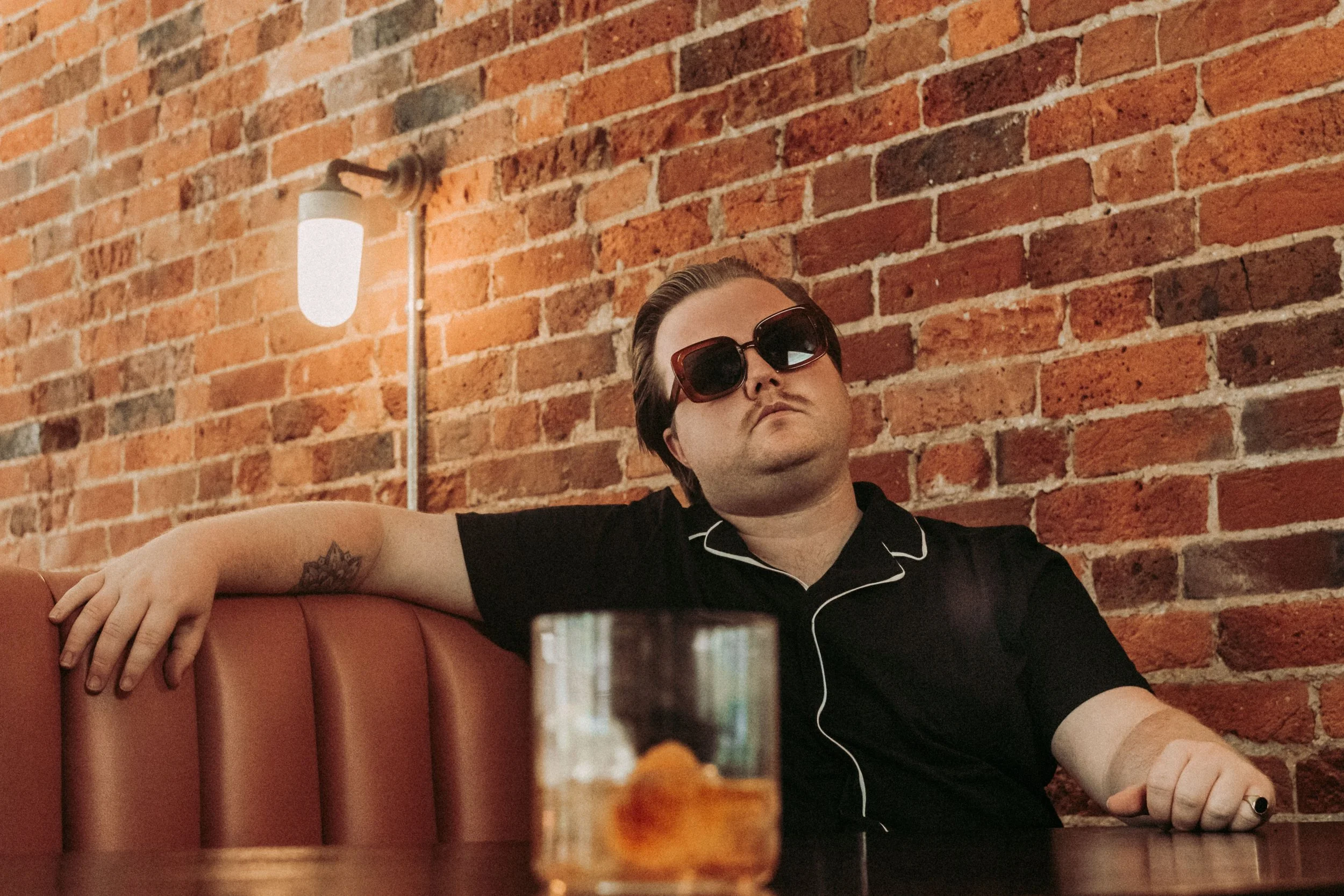 A man with dark hair and wearing large black sunglasses is sitting in a restaurant booth with a red leather seat against an exposed brick wall. He is wearing a black shirt with white piping and has a tattoo on his arm. There is a lamp on the brick wa