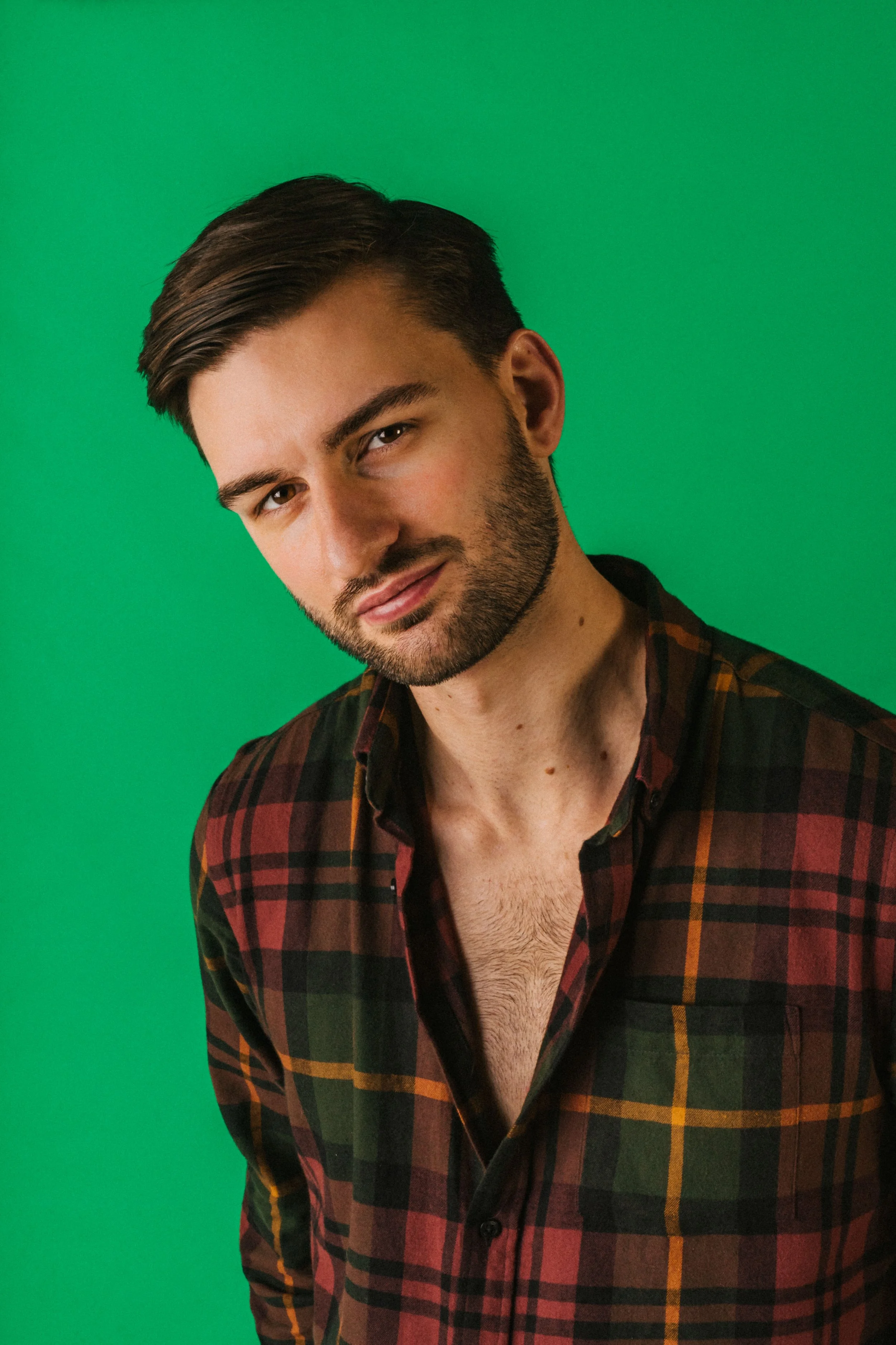 A young man with dark hair, a beard, and light skin wearing a plaid shirt partially unbuttoned against a green background.