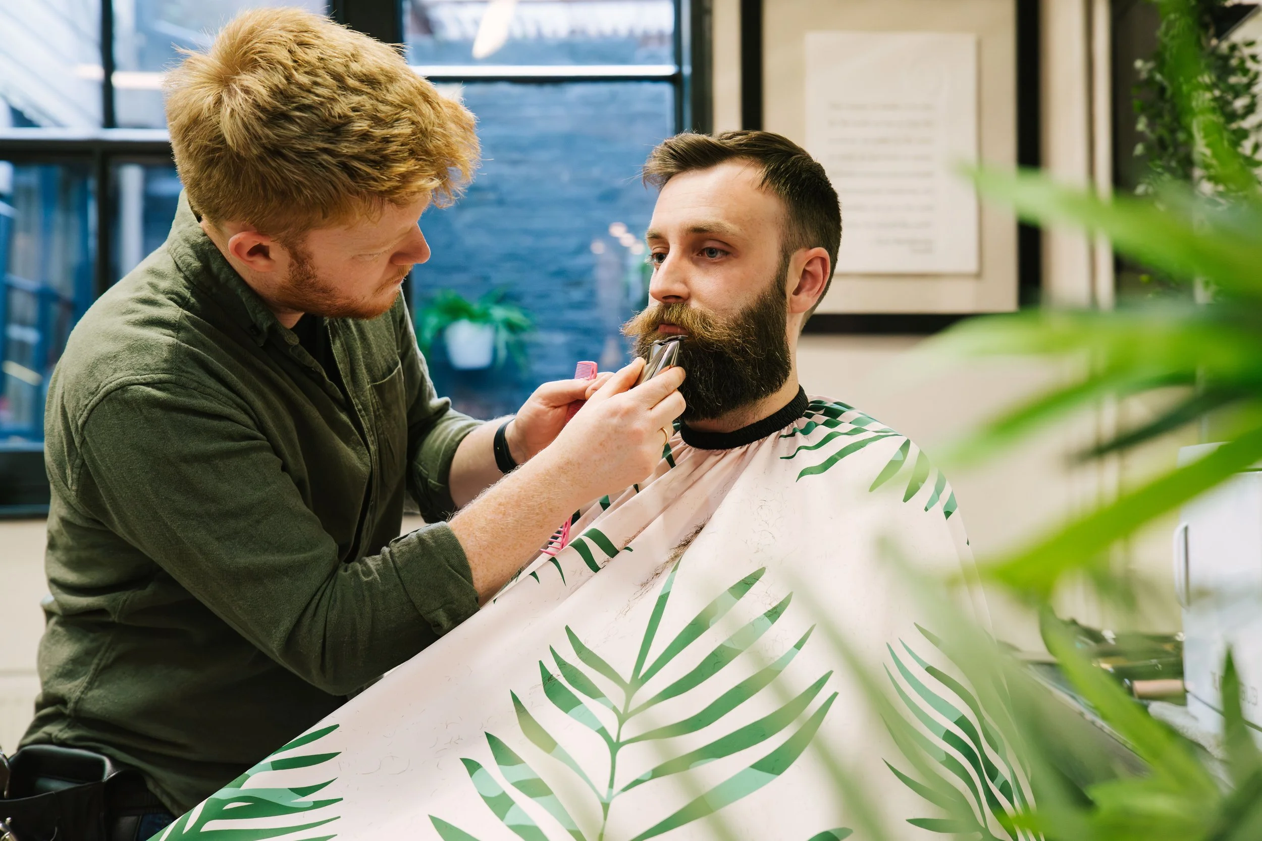 Barber trims a man's beard with electric clipper inside a barber shop, with a tropical leaf-patterned cape around the man.