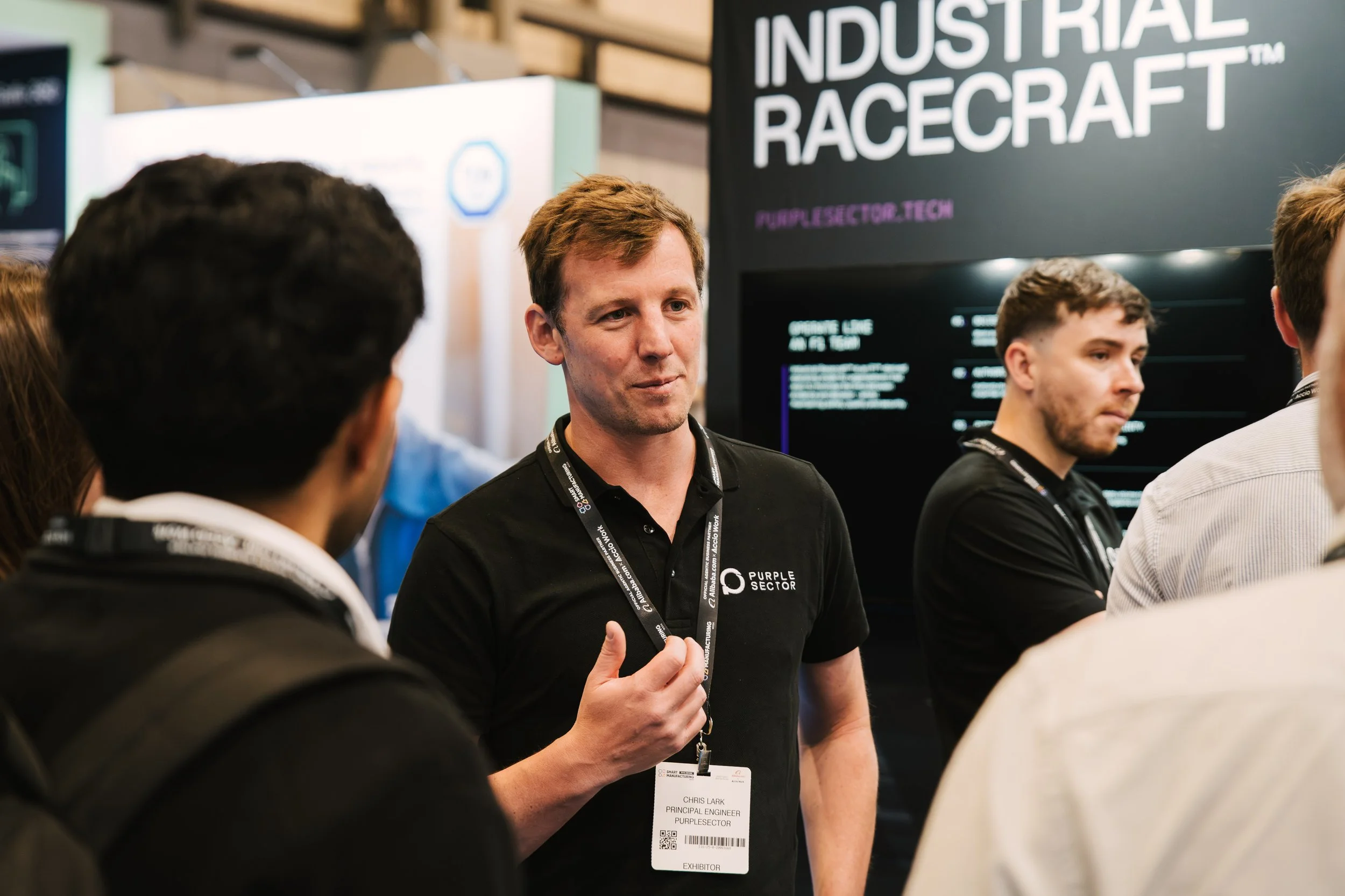 A man with a badge that reads 'Chris Lark, Principal Engineer, Purple Sector' is speaking to a group at a booth with a large sign reading 'Industrial Racecraft.' Several other people are listening, and there are display screens in the background.