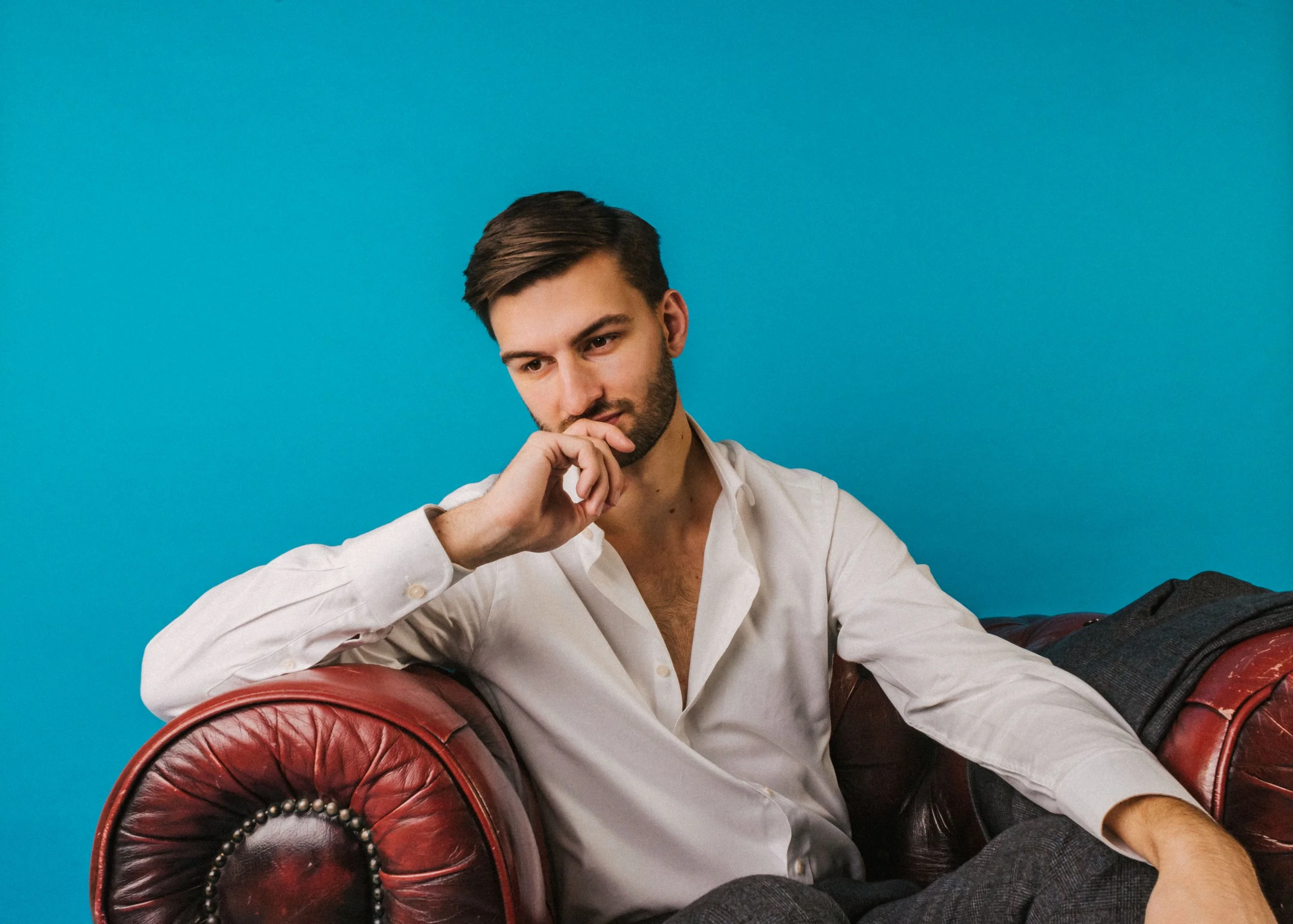 A young man with brown hair and a beard, wearing a white shirt, sitting on a red leather armchair with a blue background.