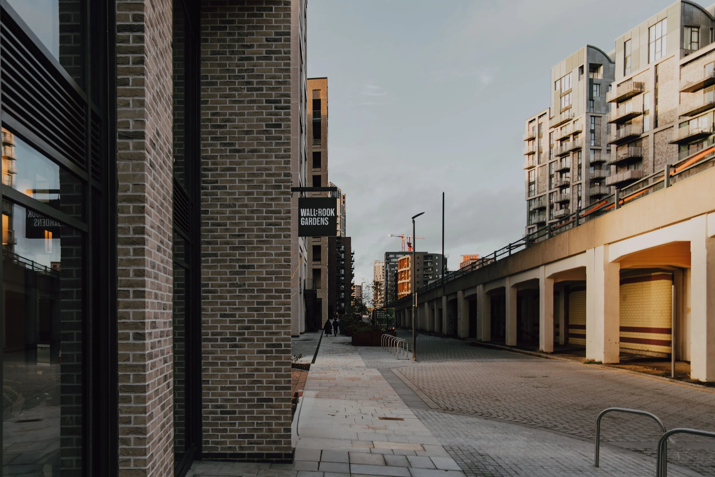 A city street view showing modern apartment buildings, a brick building, and a sidewalk with a few pedestrians walking. There's a sign that reads 'Wallbrook Gardens' attached to one of the buildings. The sky is partly cloudy.