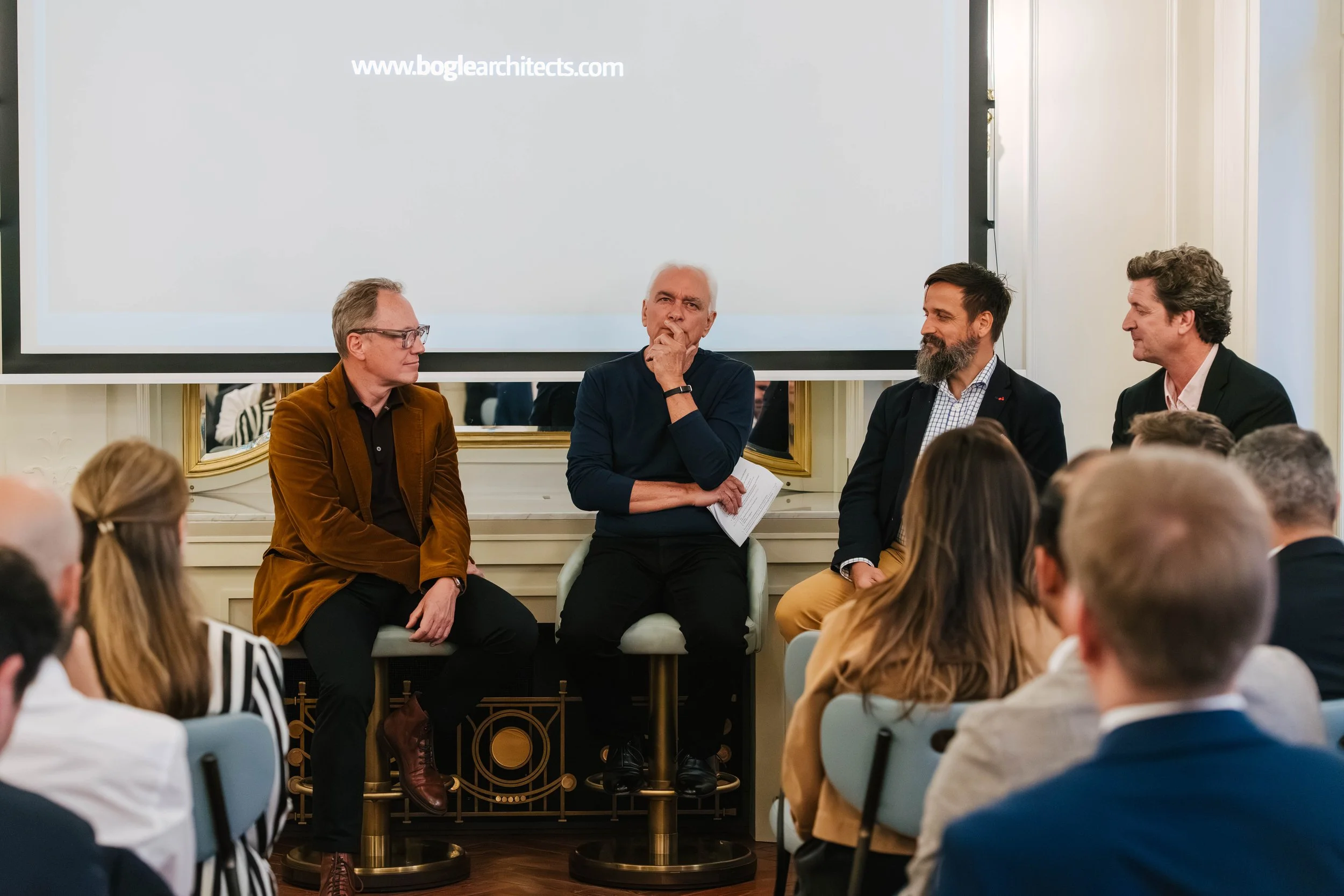 Four men seated in front of an audience during a panel discussion in a formal setting. One man is wearing glasses and an orange-brown blazer, another has white hair and is wearing a navy sweater, the third has a beard and dark hair with a checked shi