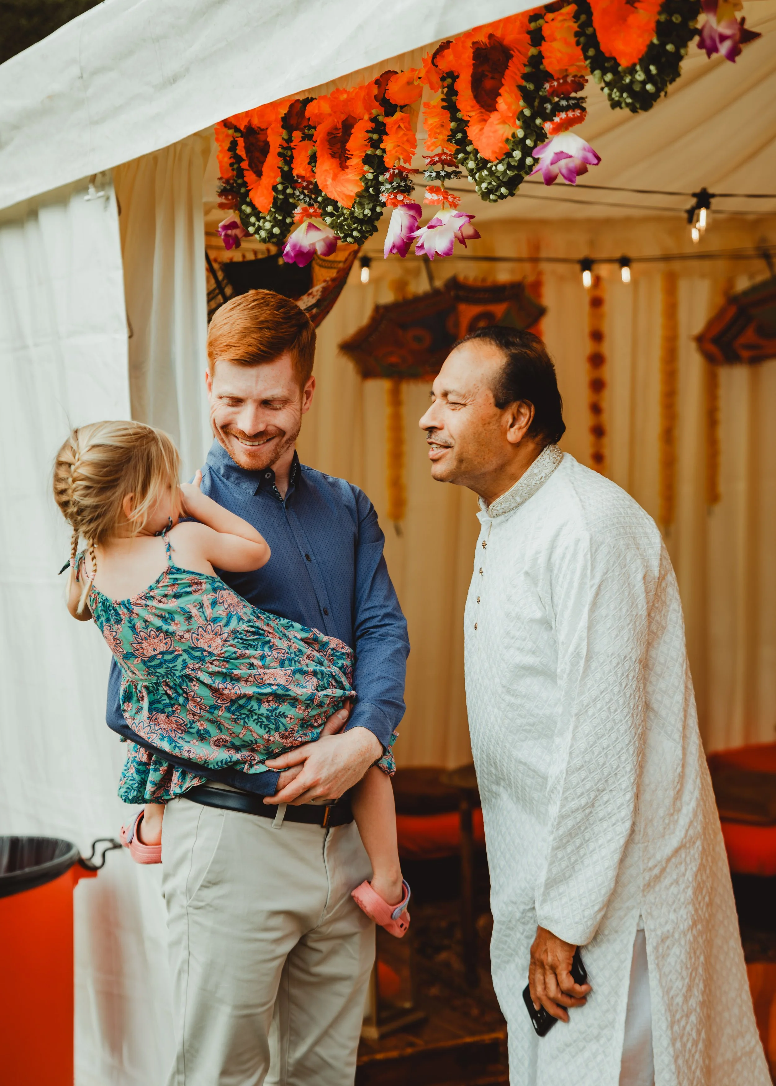 Three men and a young girl smiling and talking inside a decorated tent at a celebration, with colorful garlands and lanterns hanging from the ceiling.
