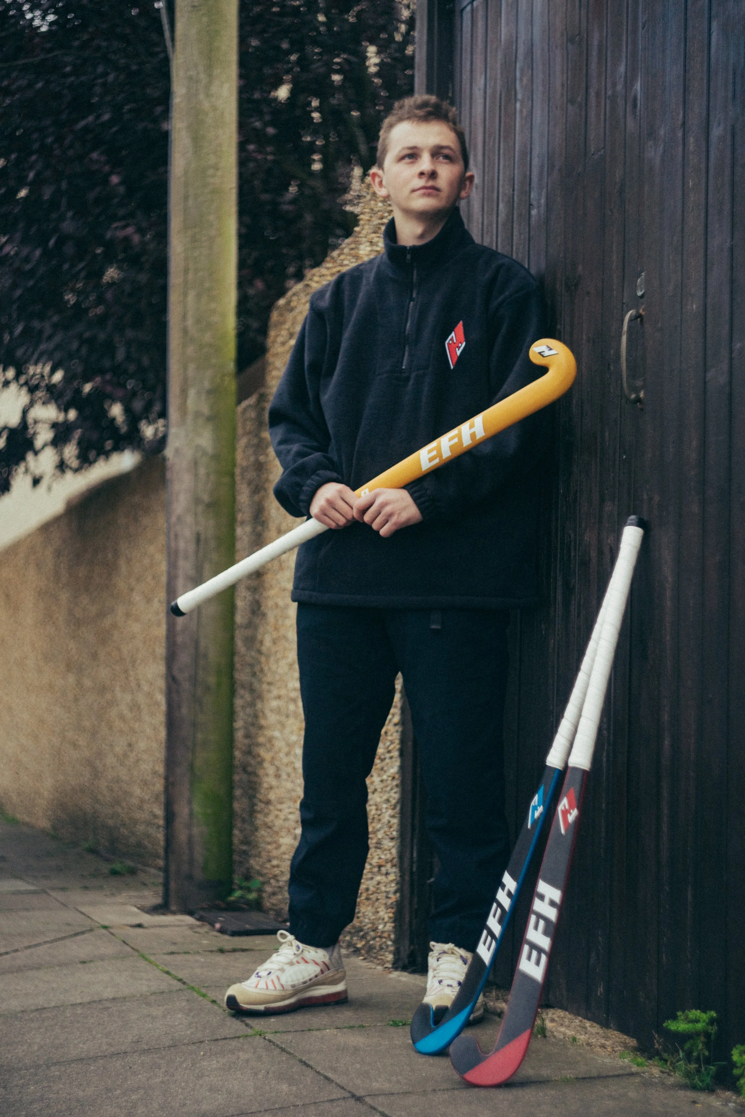 A young man standing outdoors against a dark wooden fence, holding a yellow hockey stick with the label 'EHH,' with a pair of hockey sticks on the ground nearby, wearing a black fleece jacket, black pants, and white sneakers.