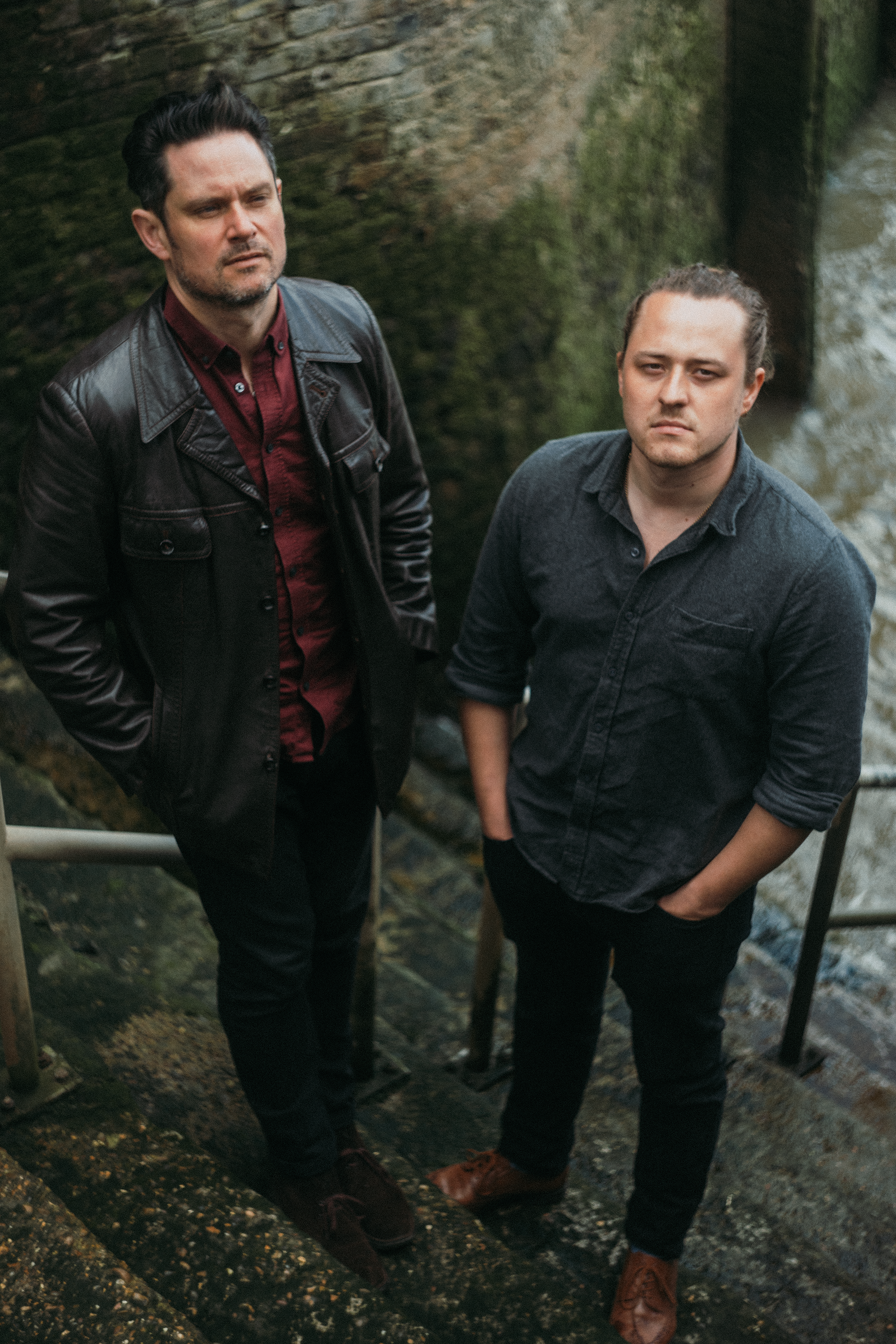 Two men standing on a stone staircase beside a moss-covered rock wall, facing the camera with serious expressions.